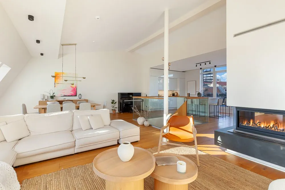 Modernes Wohnzimmer mit Kamin, weißen Sofas, Holztischen und offener Küche im Hintergrund.