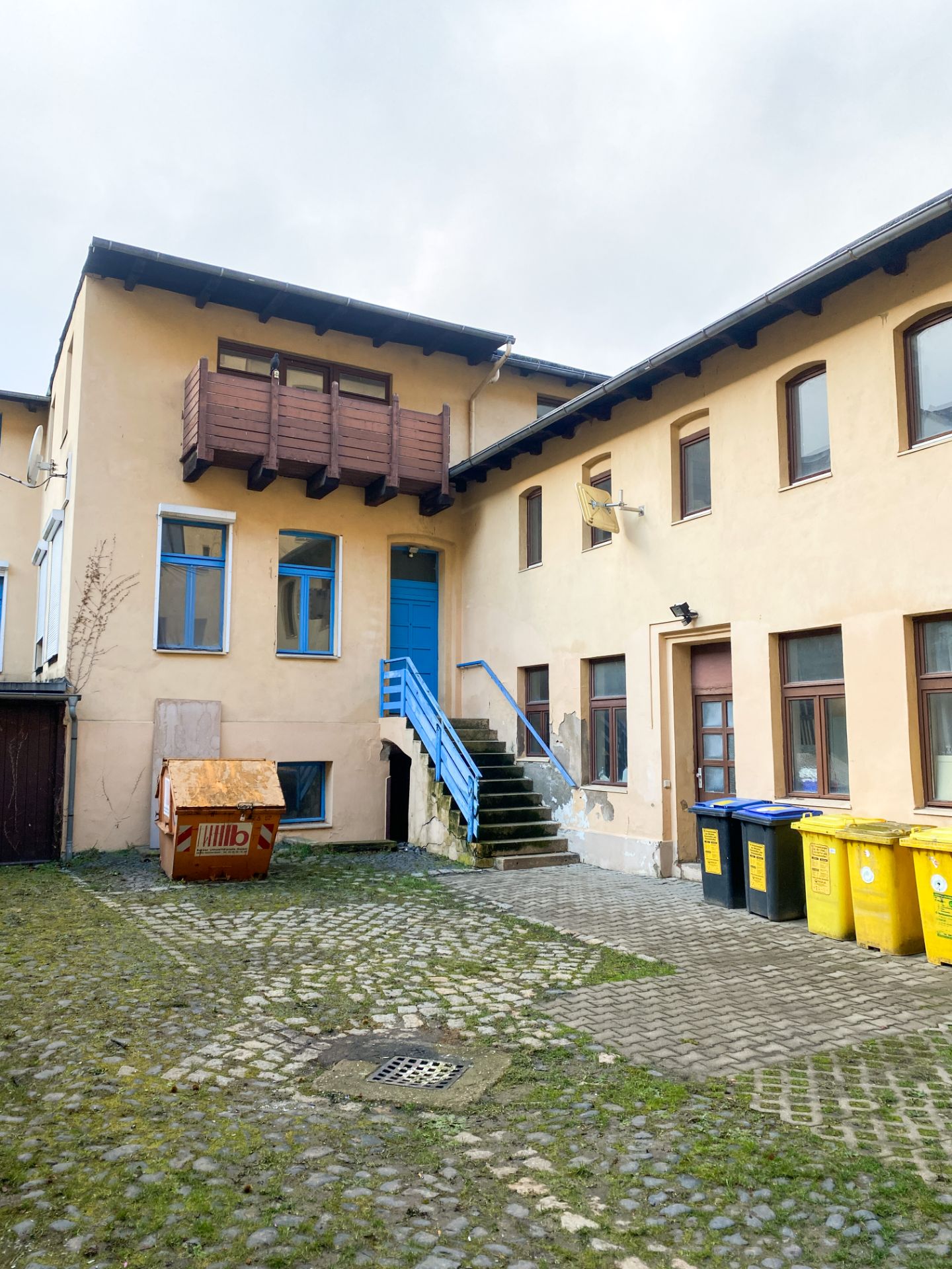 Innenhof mit gelbem Gebäude, blauen Treppen, Balkon, Mülltonnen und Abfallcontainer.