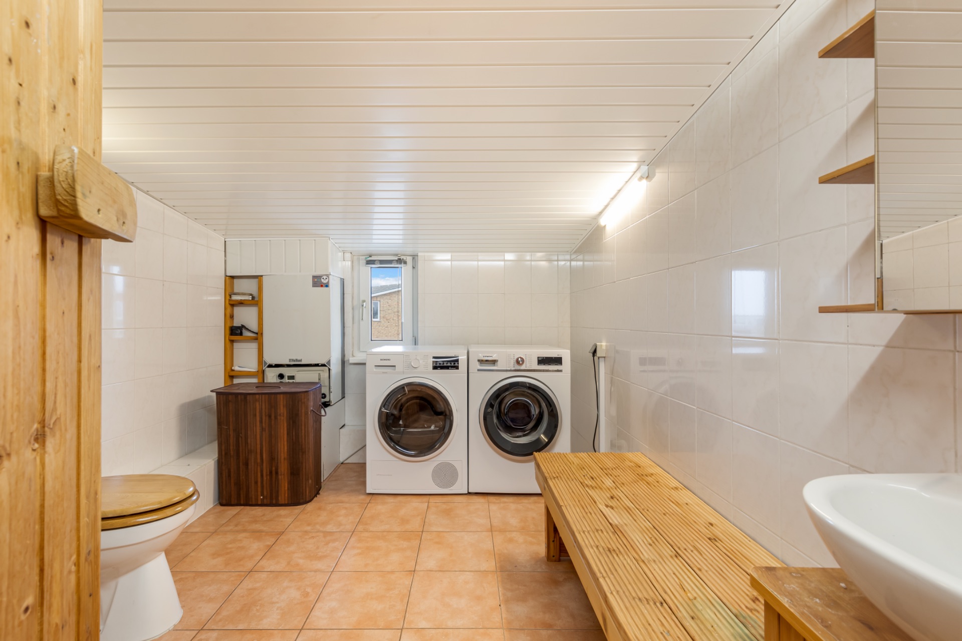 Hauswirtschaftsraum mit Waschmaschine, Trockner, Holzbank, Waschbecken und Toilette.