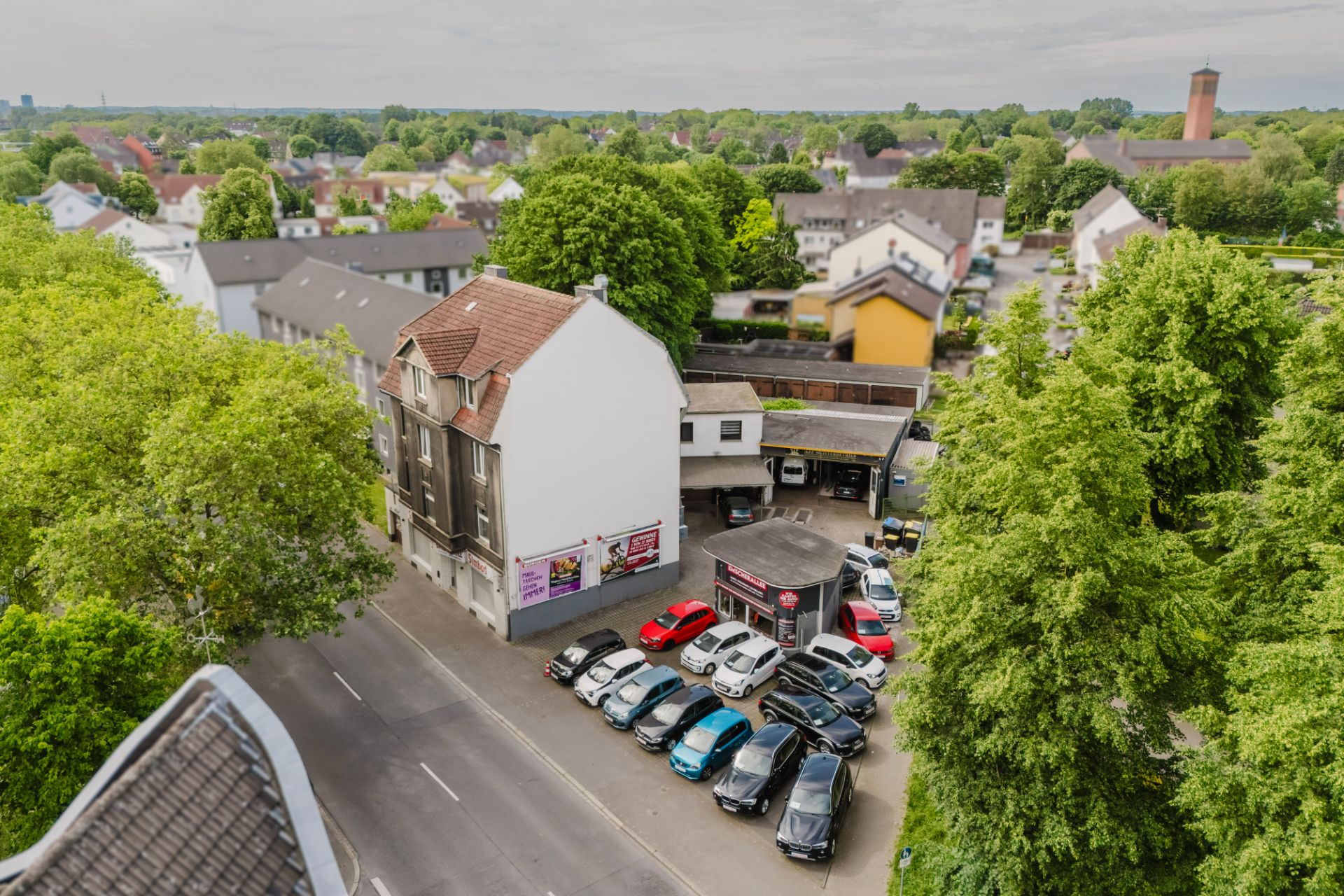Luftaufnahme von Wohnhäusern, einer Straße und einem kleinen Parkplatz mit vielen Autos, umgeben von Bäumen.