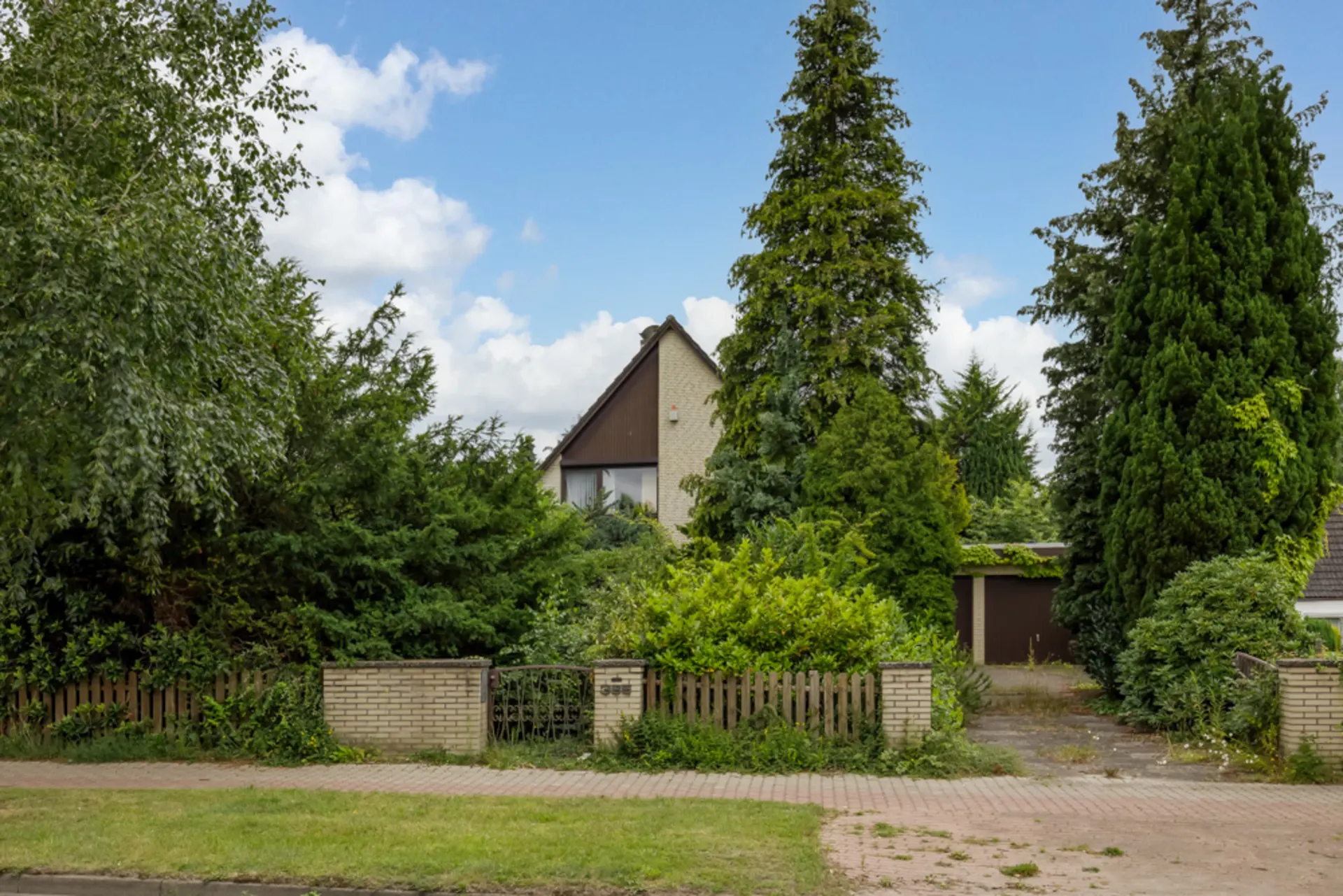 Einfamilienhaus mit Satteldach hinter vielen Bäumen und Sträuchern, Einfahrt und Tor sichtbar.