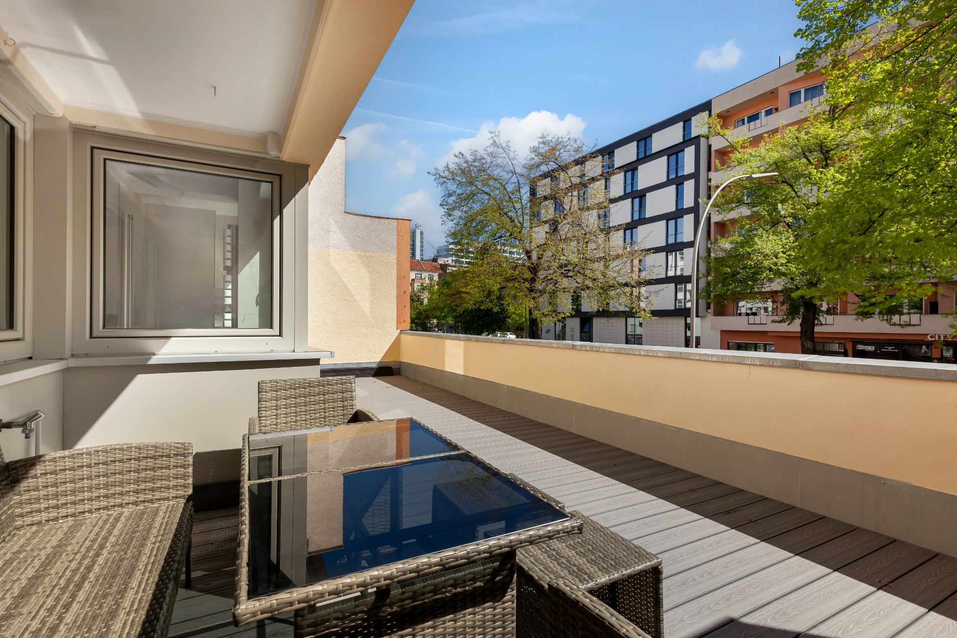 Große Terrasse mit Glas-Tisch und Stühlen, Blick auf Wohngebäude und Bäume.