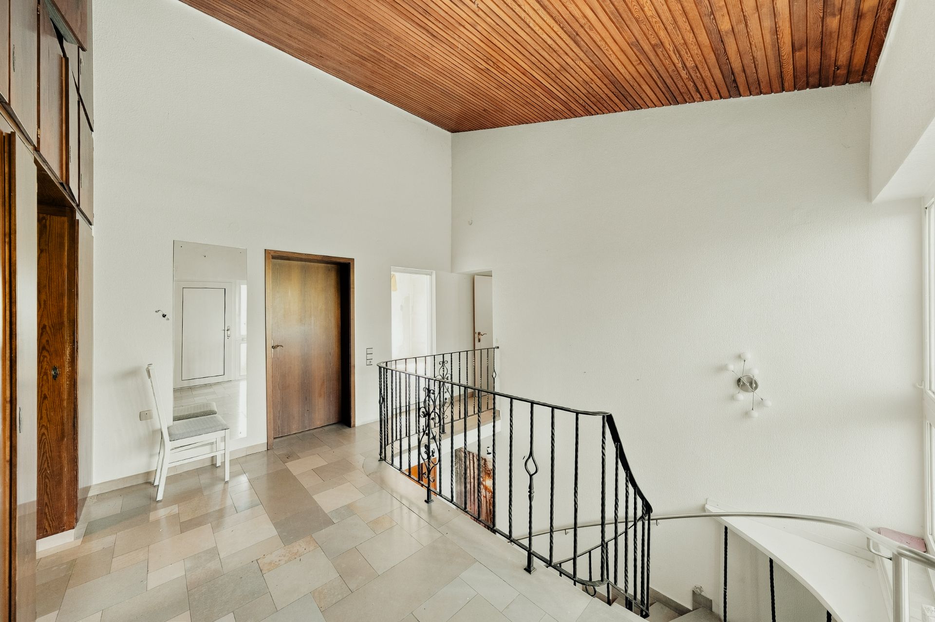 Heller Flur mit Treppe, weißen Wänden, Holztür und Holzdecke, Stuhl an der Wand.