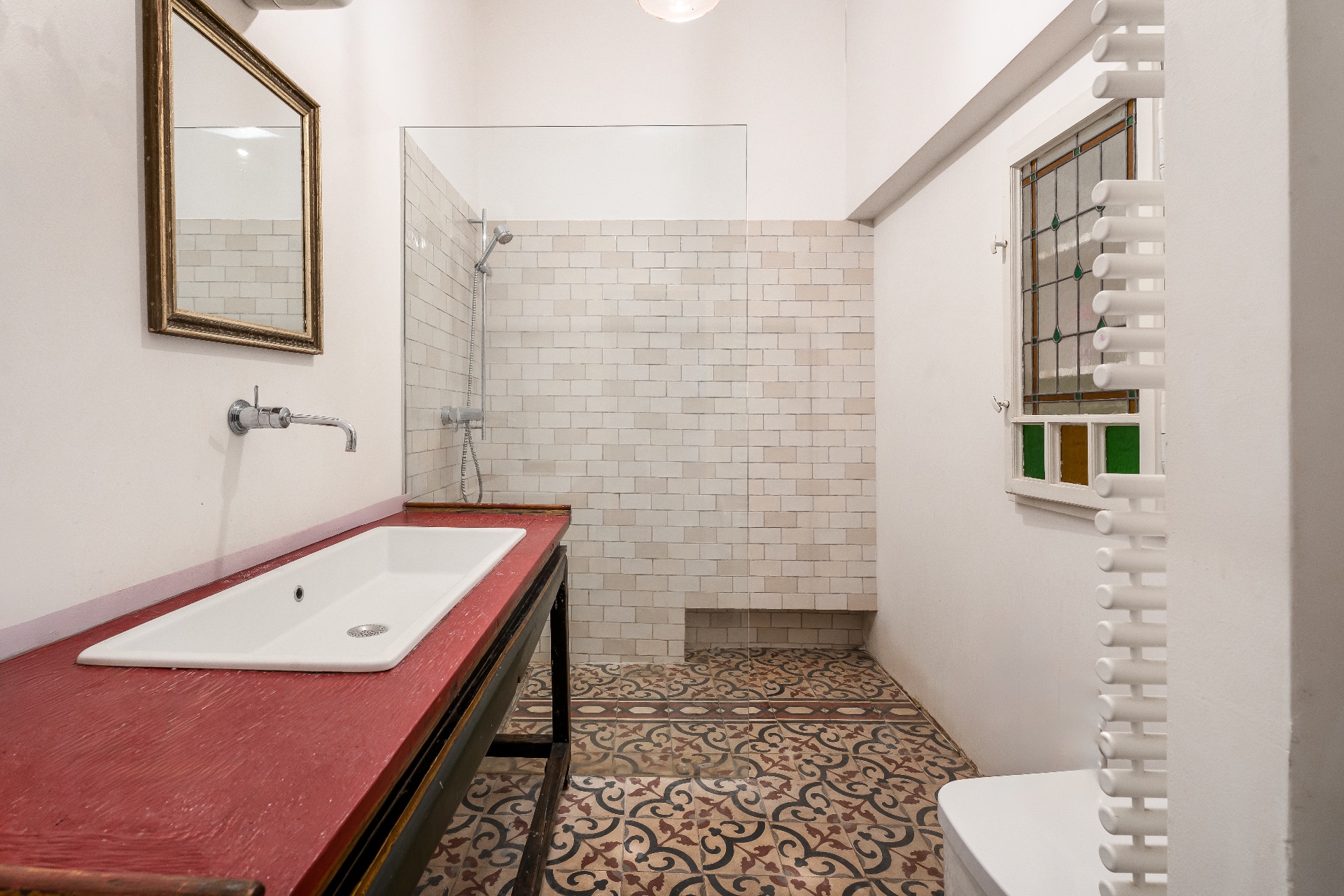 Helles Badezimmer mit gemustertem Fliesenboden, großer Dusche und roter Waschtischplatte.