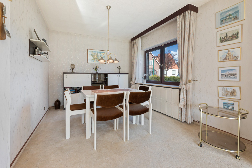 Esszimmer mit Tisch, sechs Stühlen, Fenster, Kommode, Wandbildern und Teewagen.