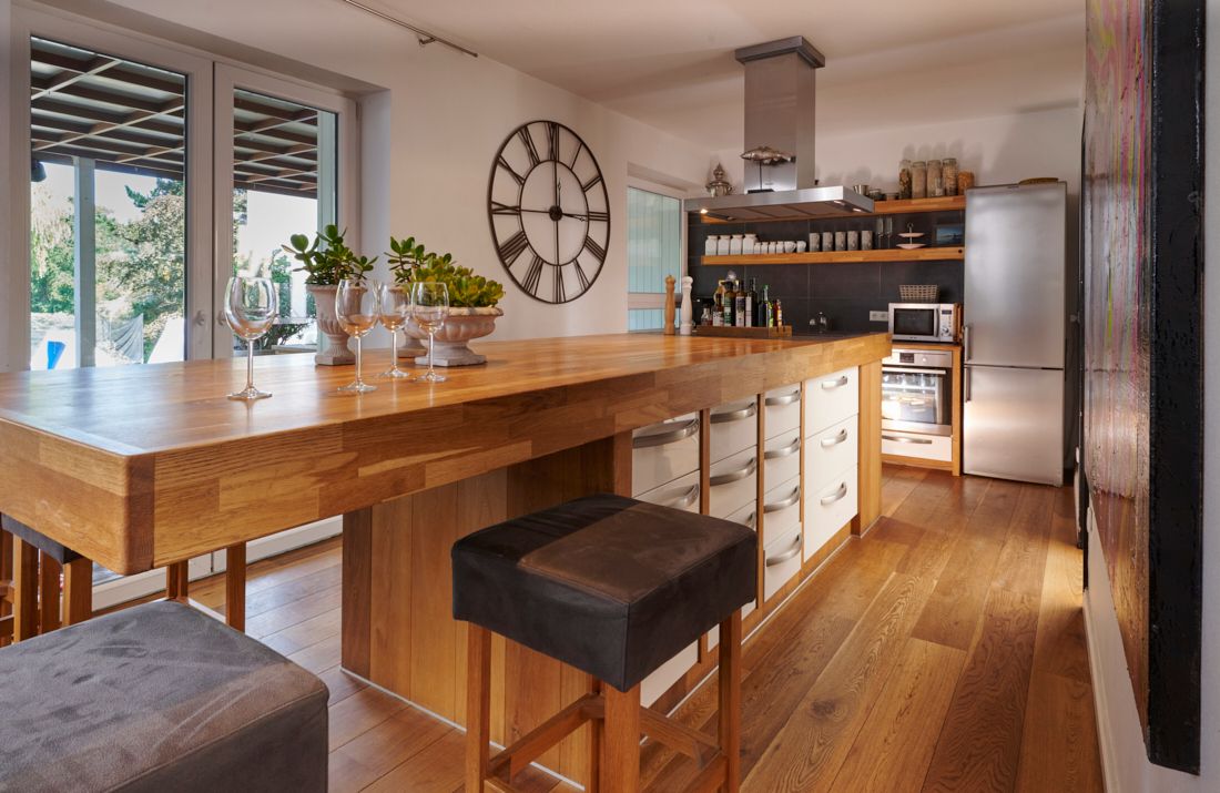 Moderne Küche mit großem Holztisch, Barhockern und Wanduhr, daneben Fenster und Kühlschrank.