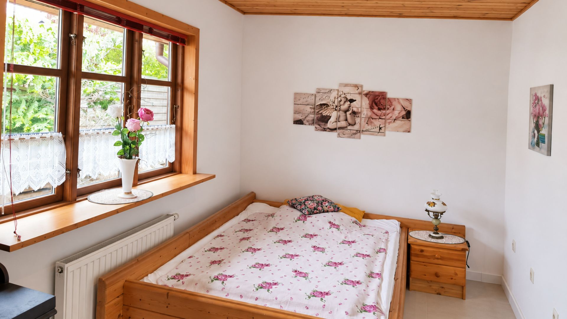 Kleines Schlafzimmer mit Holzbett, Blumenbettwäsche, Fensterbank mit Blume und Nachttischlampe.