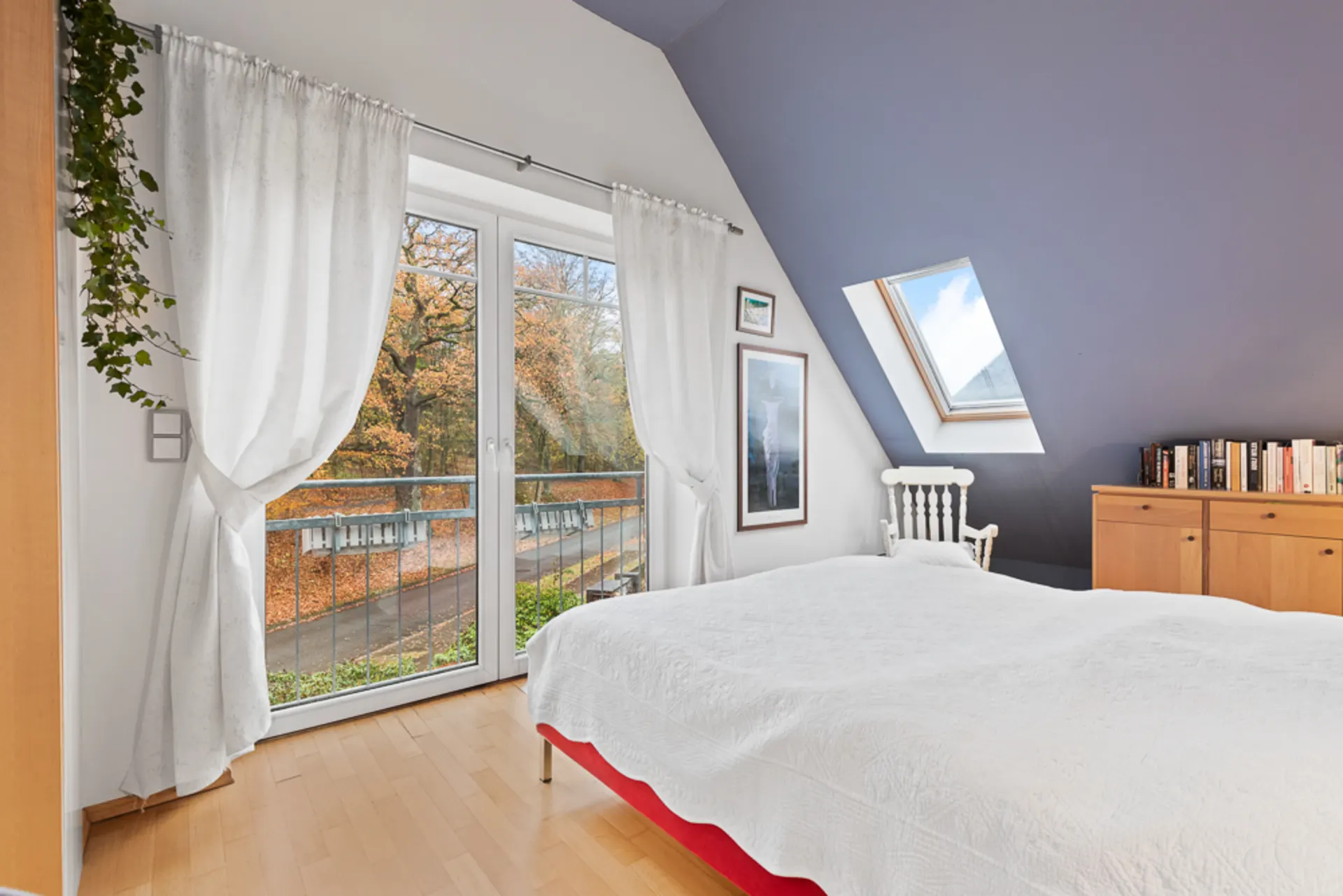 Helles Schlafzimmer mit großem Fenster, Vorhängen, Dachfenster, Bett und Kommode mit Büchern.