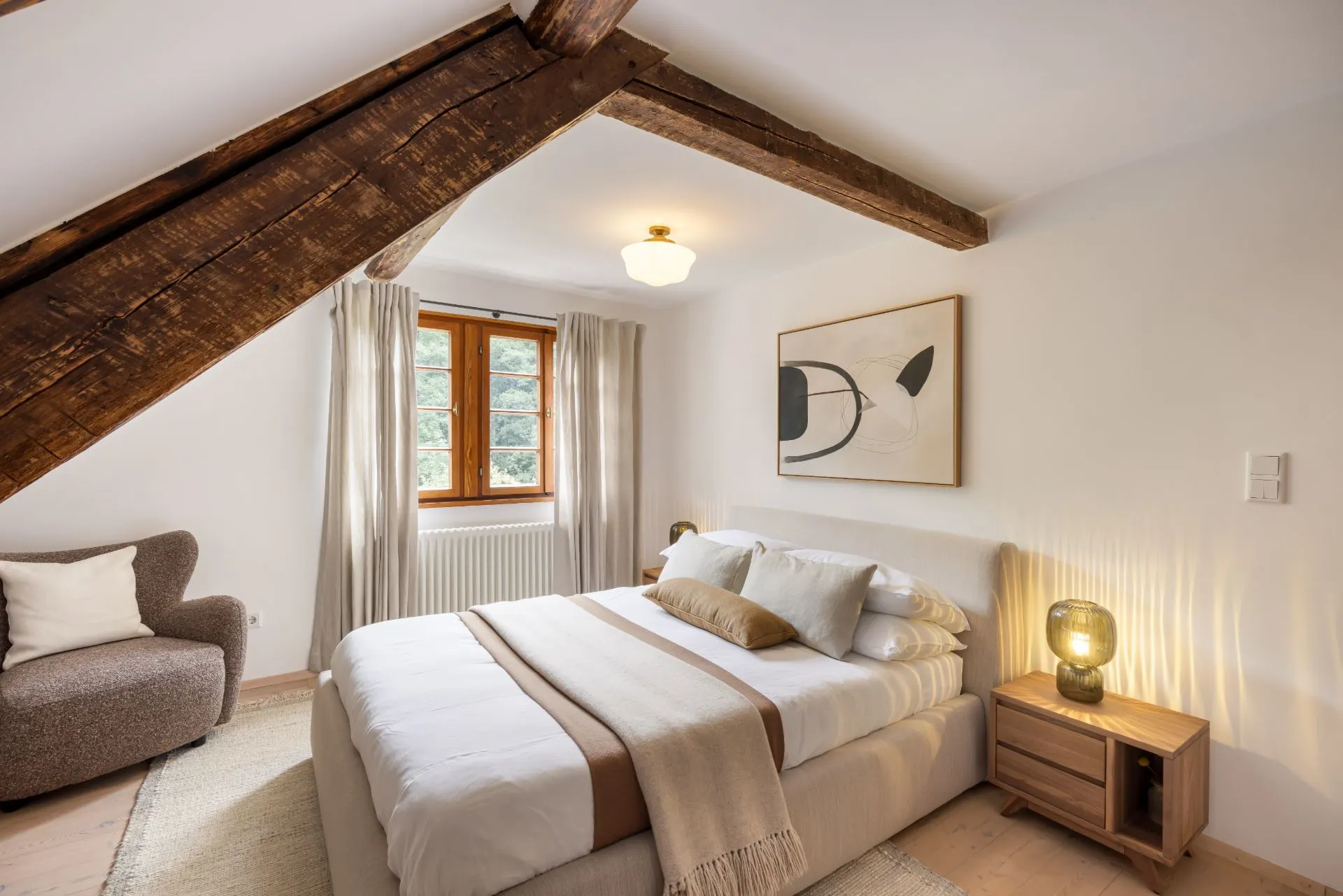 Helles Schlafzimmer mit Holzbalken, Doppelbett, Fenster und Nachttischlampe.