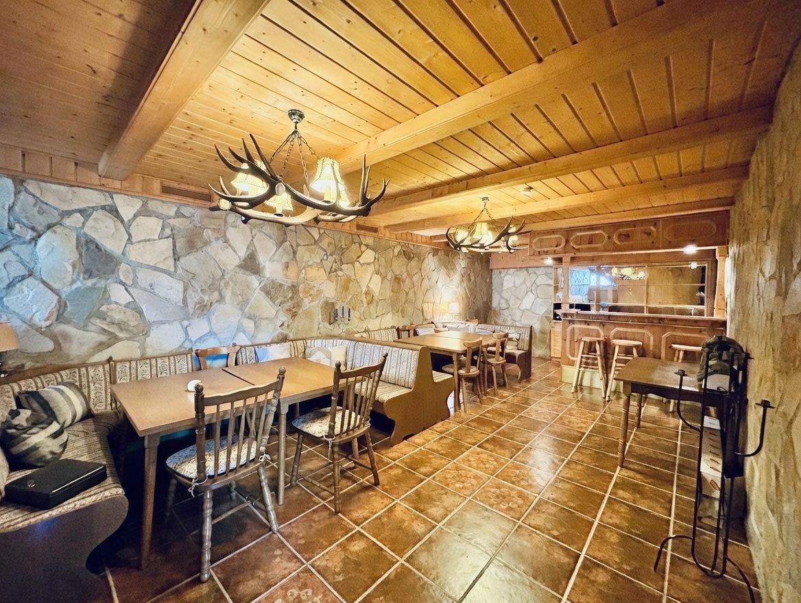 Rustikaler Essbereich mit Holzdecke, Steinwänden, Holztischen, Bänken und Geweihlampen.