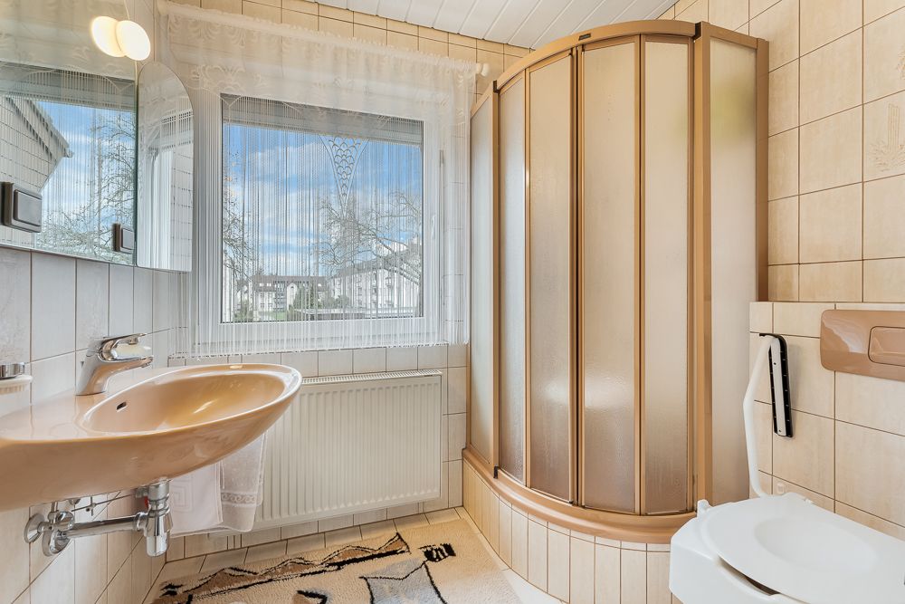 Helles Badezimmer mit Dusche, Waschbecken, Toilette und großem Fenster mit Blick nach draußen.