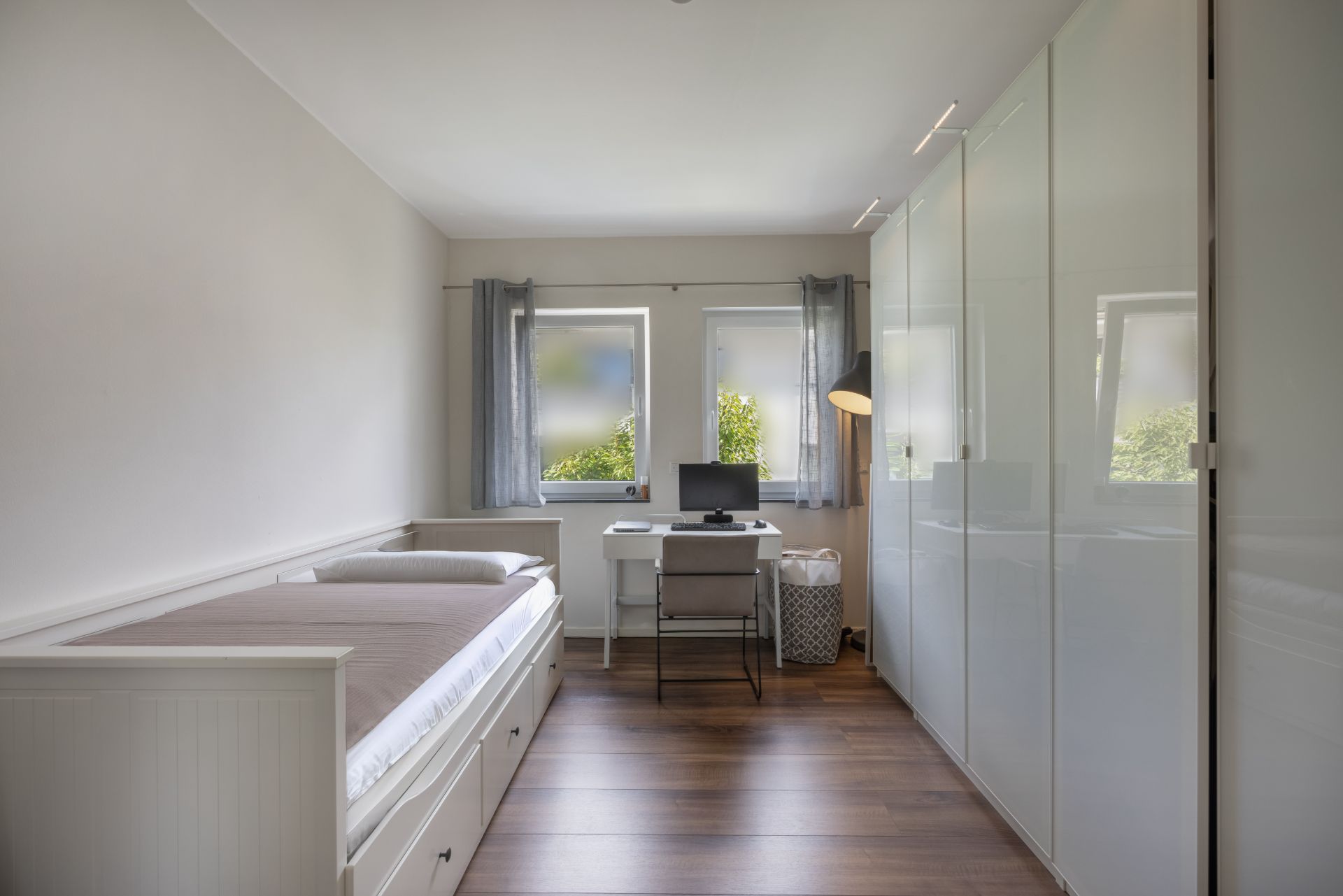 Schlafzimmer mit Einzelbett, Schreibtisch, Computer und großem weißen Kleiderschrank.