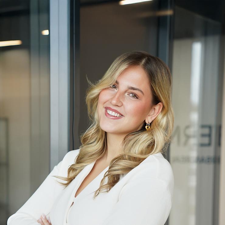 Junge Frau mit langen blonden Haaren und weißem Oberteil steht lächelnd mit verschränkten Armen.