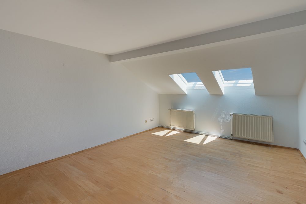 Leerer Raum mit Holzfußboden, zwei Dachfenstern und zwei Heizkörpern an der Wand.
