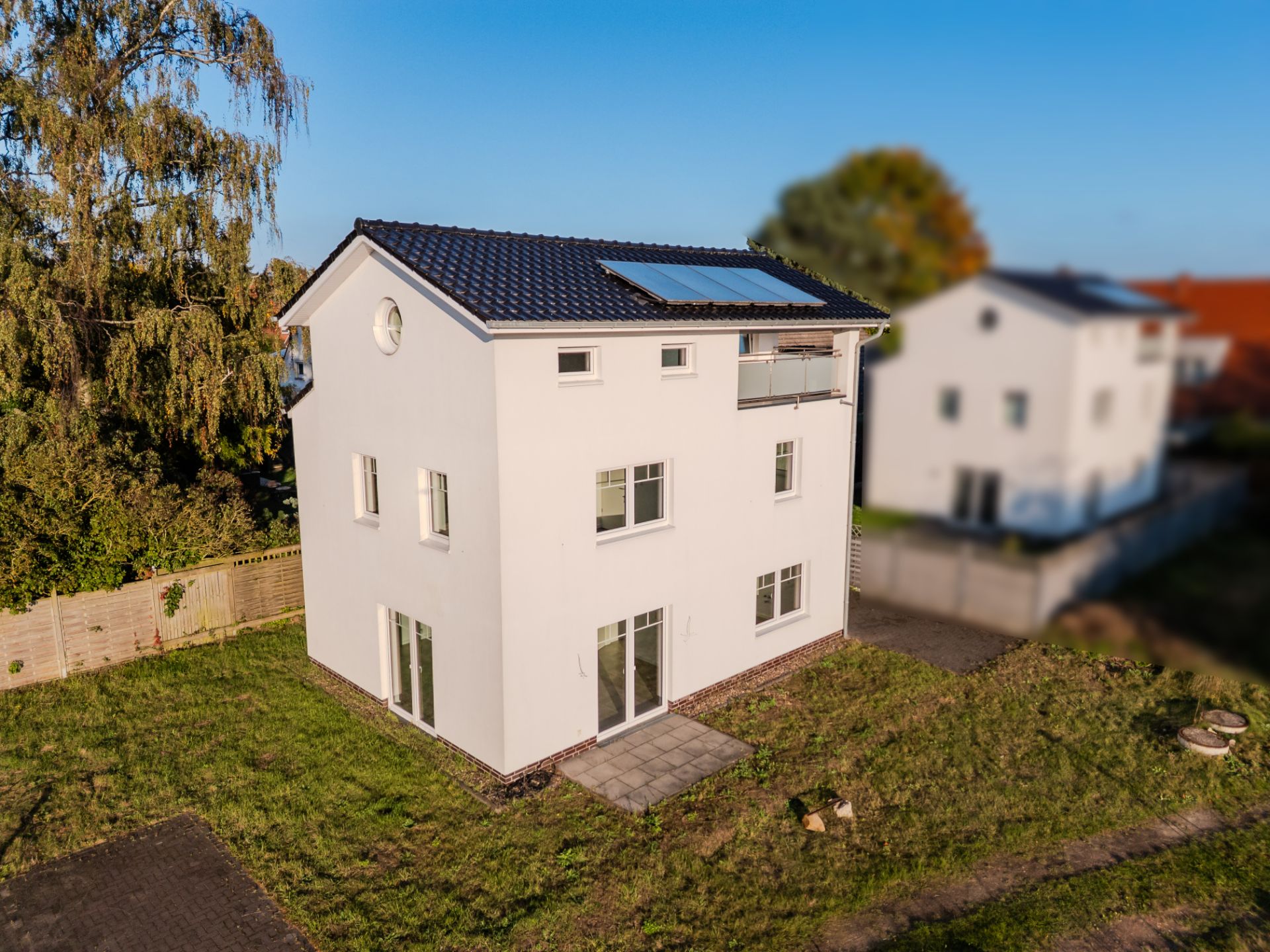 Modernes, weißes Einfamilienhaus mit dunklem Dach, Balkon und Solarpanel im Garten.