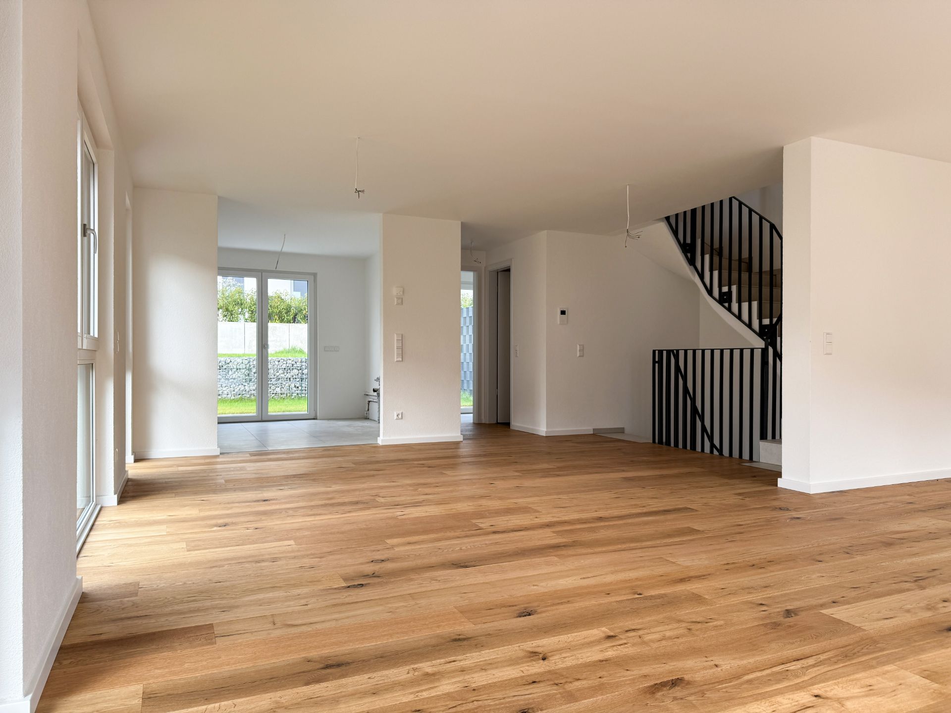 Helles Wohnzimmer mit Parkettboden, großen Fenstern und einer Treppe mit schwarzem Geländer.