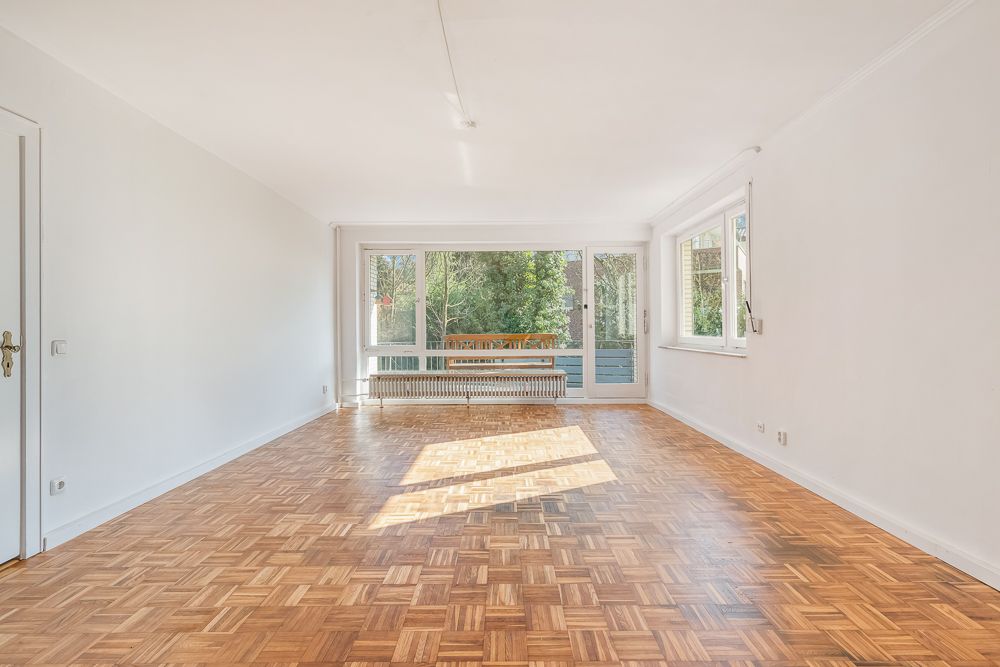 Heller leerer Raum mit Parkettboden, großen Fenstern und Blick ins Grüne.