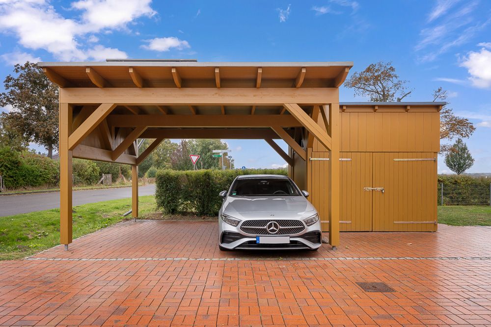Auto steht unter einem Holzcarport auf rotem Pflaster neben einem Holzschuppen im Freien.