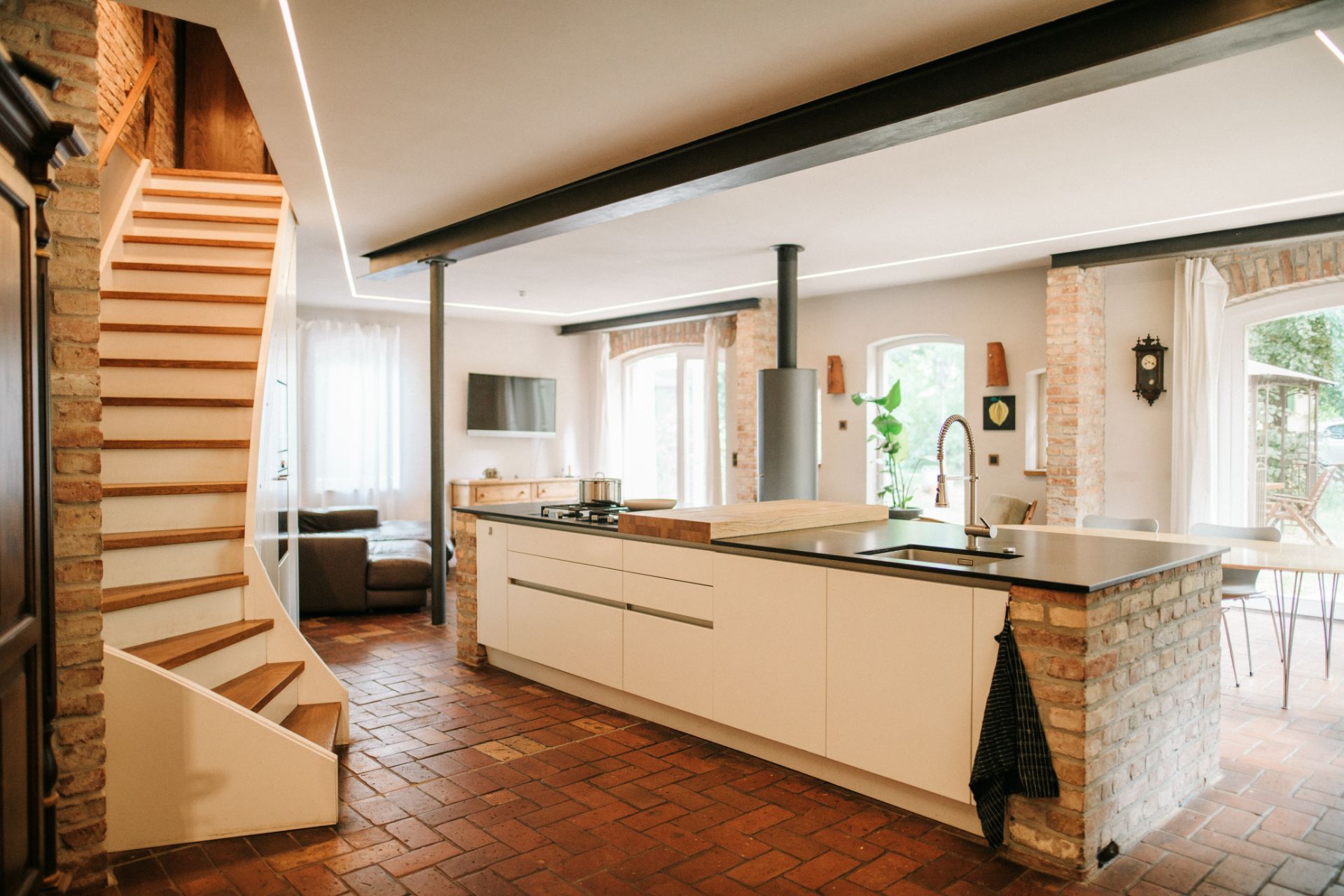 Moderne offene Küche mit Kochinsel, Holztreppe und Sitzbereich in einem hellen Raum mit Backsteinwänden.