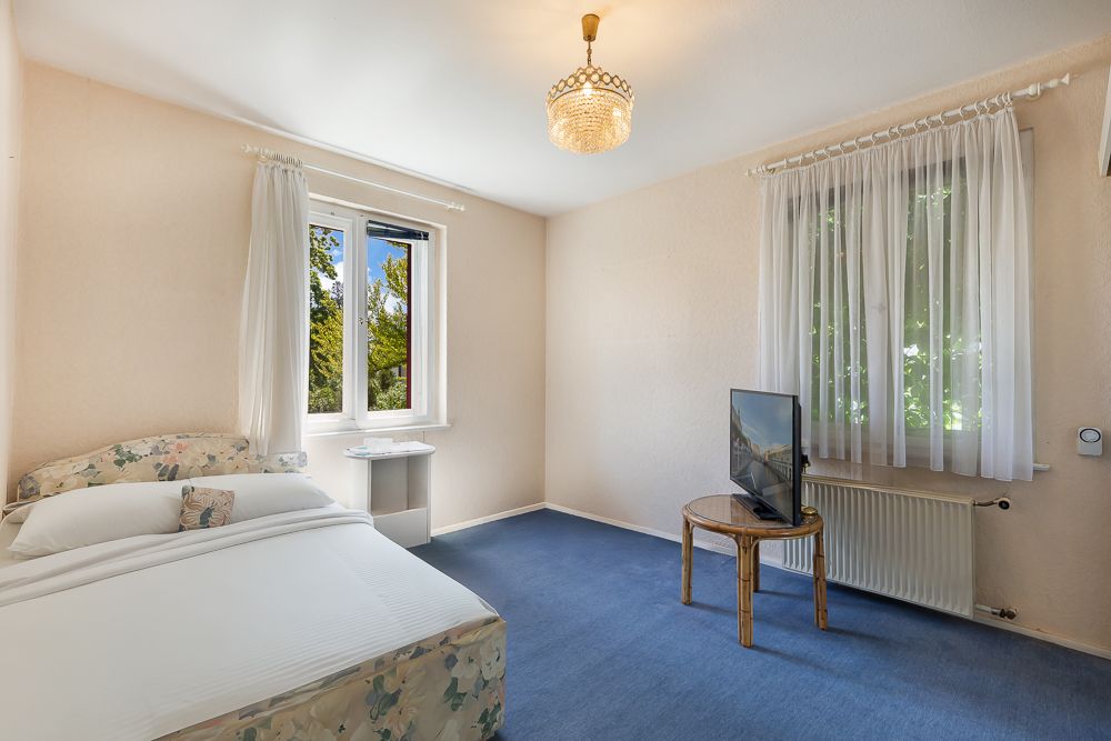 Schlafzimmer mit Doppelbett, Fernseher auf Holztisch, zwei Fenstern und blauem Teppichboden.