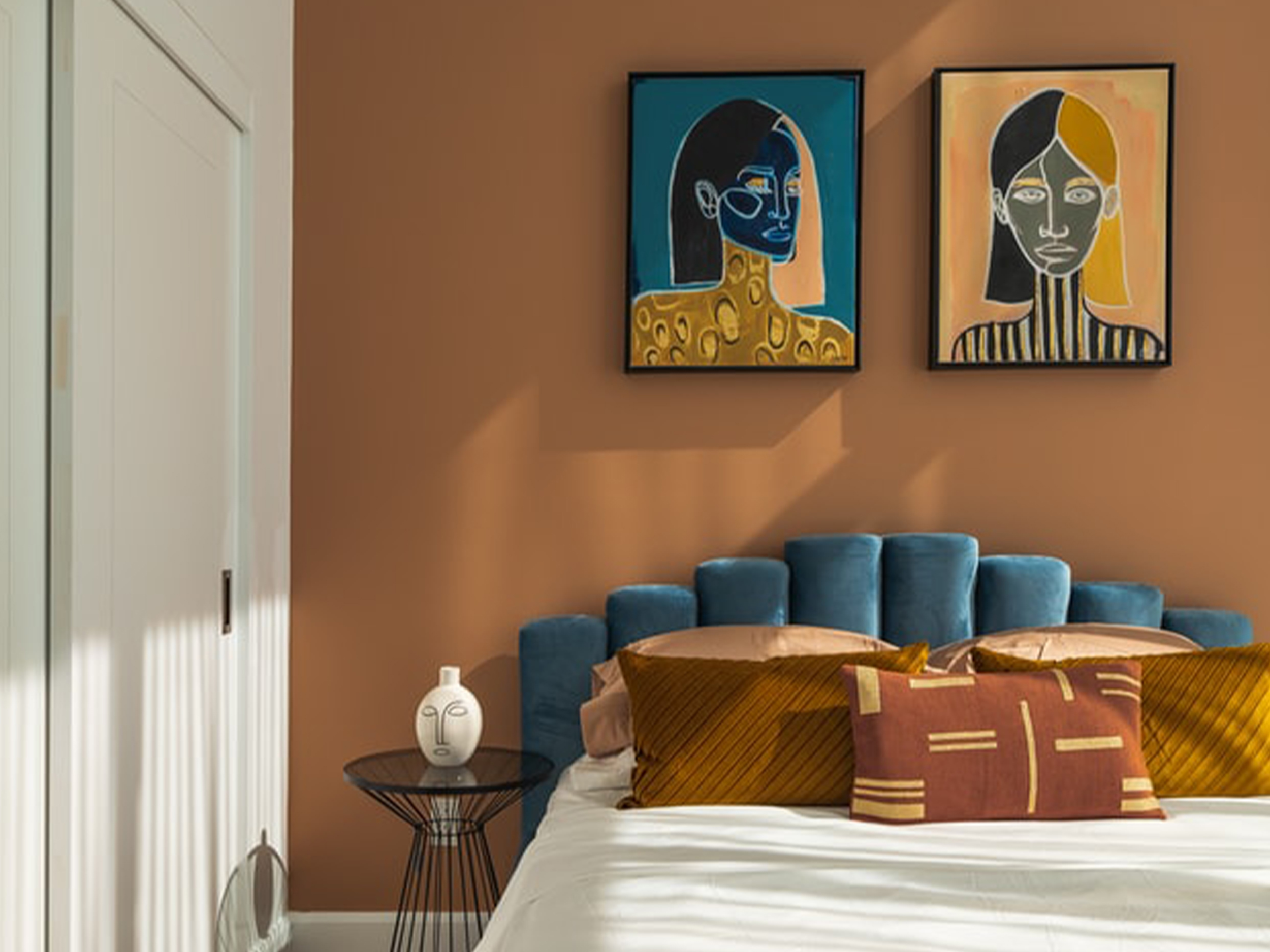 Modernes Schlafzimmer mit blauem Bett, braunen Kissen und zwei bunten Porträts an der Wand.