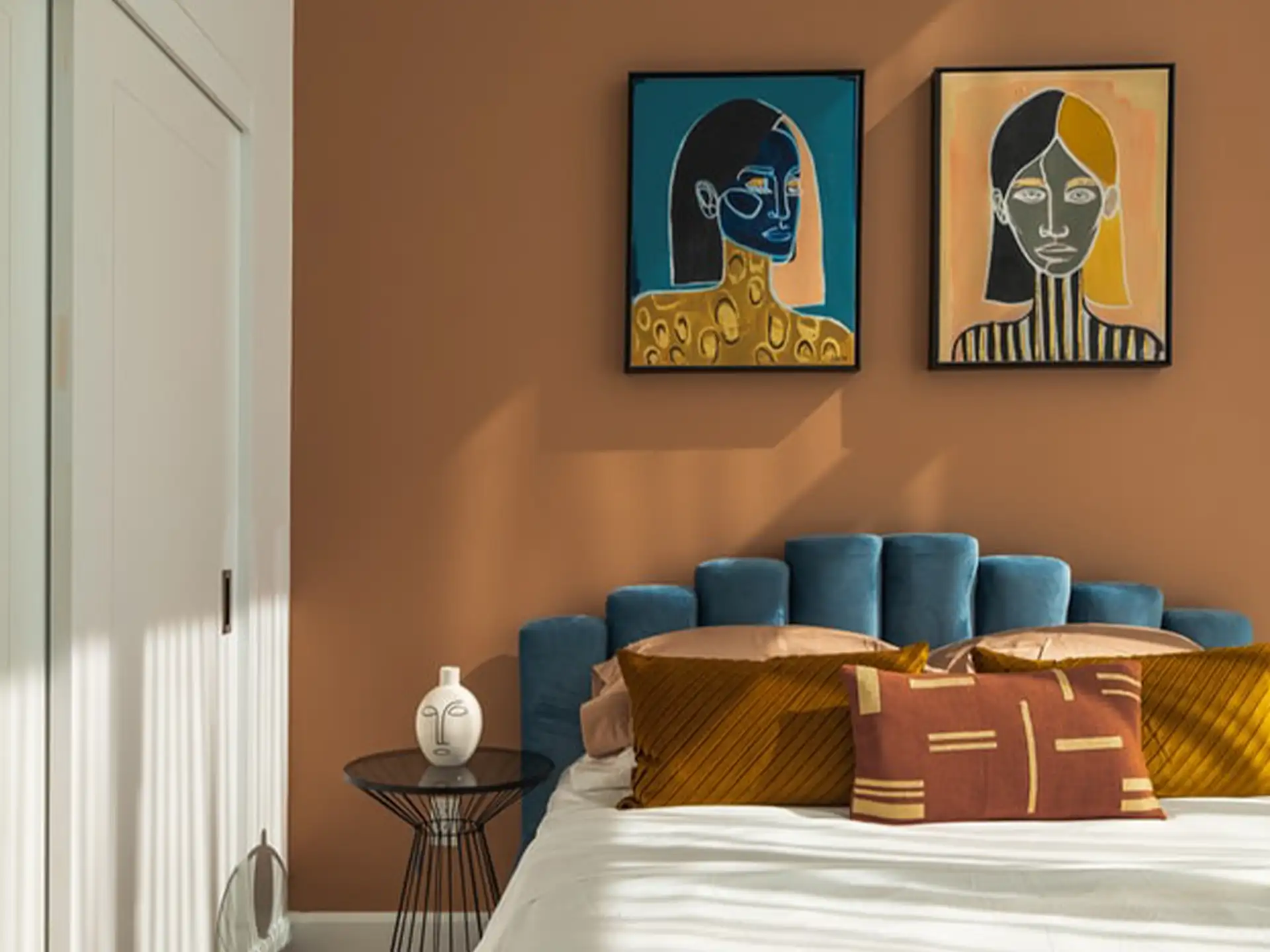 Modernes Schlafzimmer mit blauem Bett, braunen Kissen und zwei bunten Porträts an der Wand.