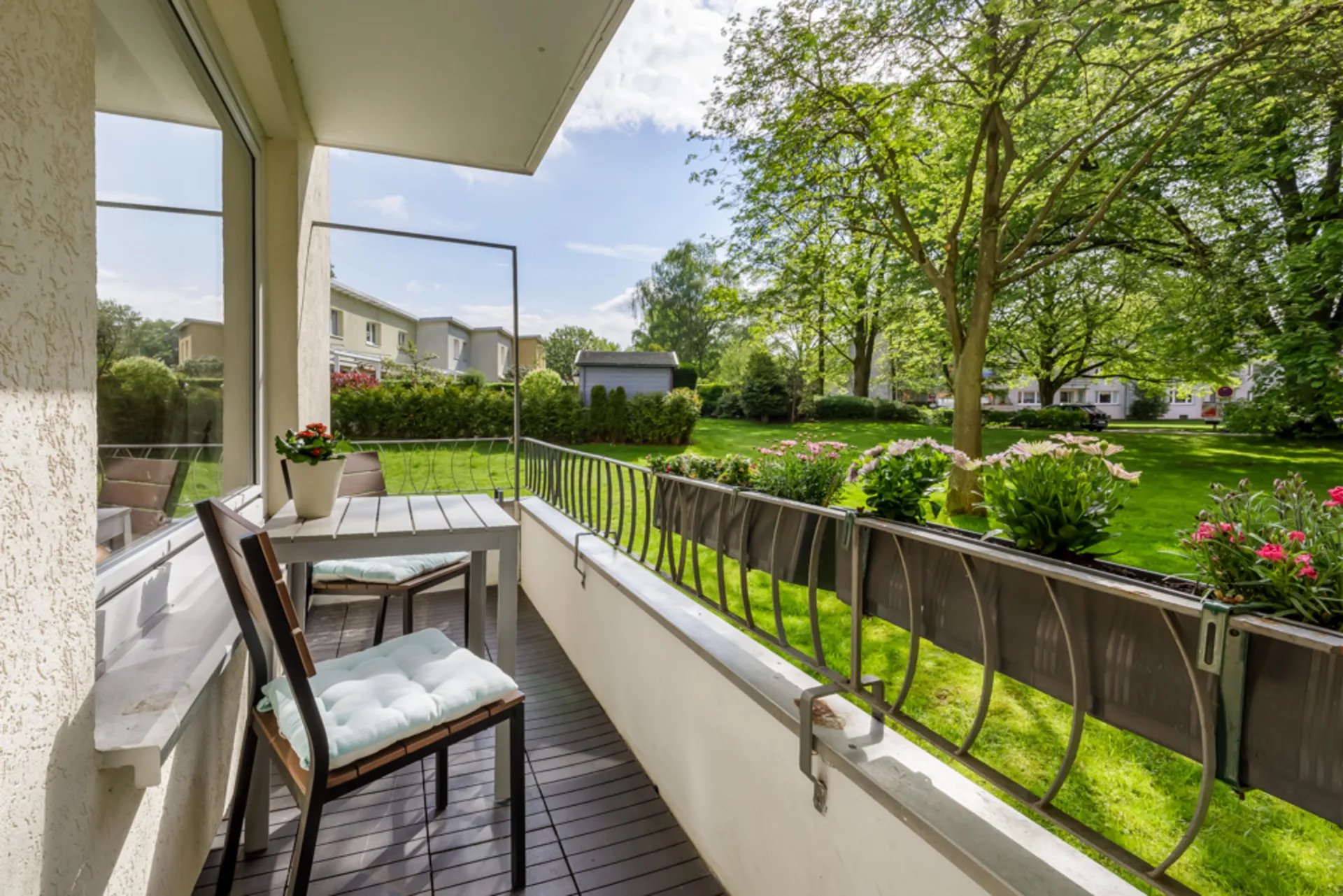 Balkon mit Tisch, zwei Stühlen und Blumenkästen, Blick auf einen grünen Garten mit Bäumen.