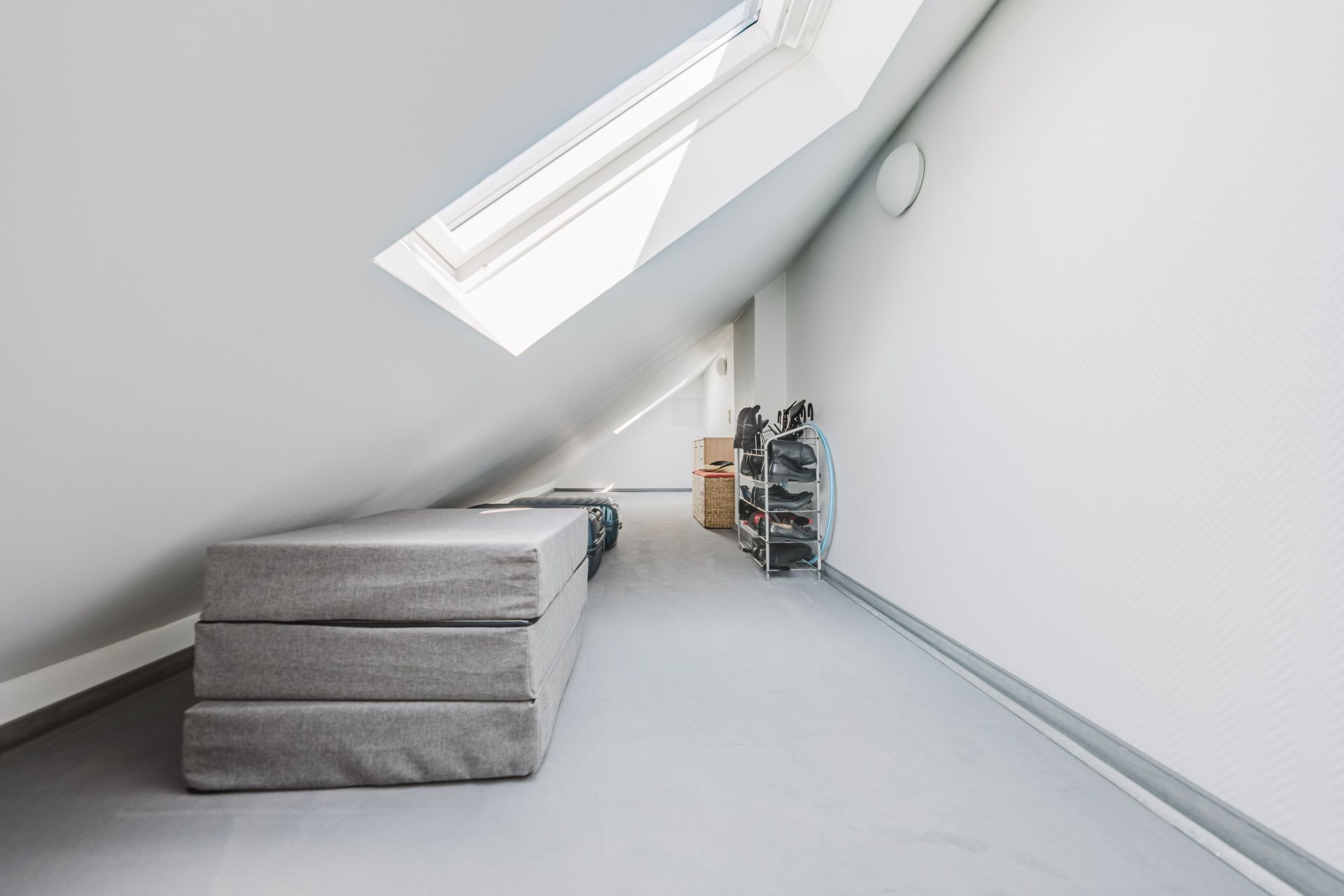 Dachzimmer mit Dachfenster, grauem Klappsofa und Schuhregal auf grauem Teppichboden.