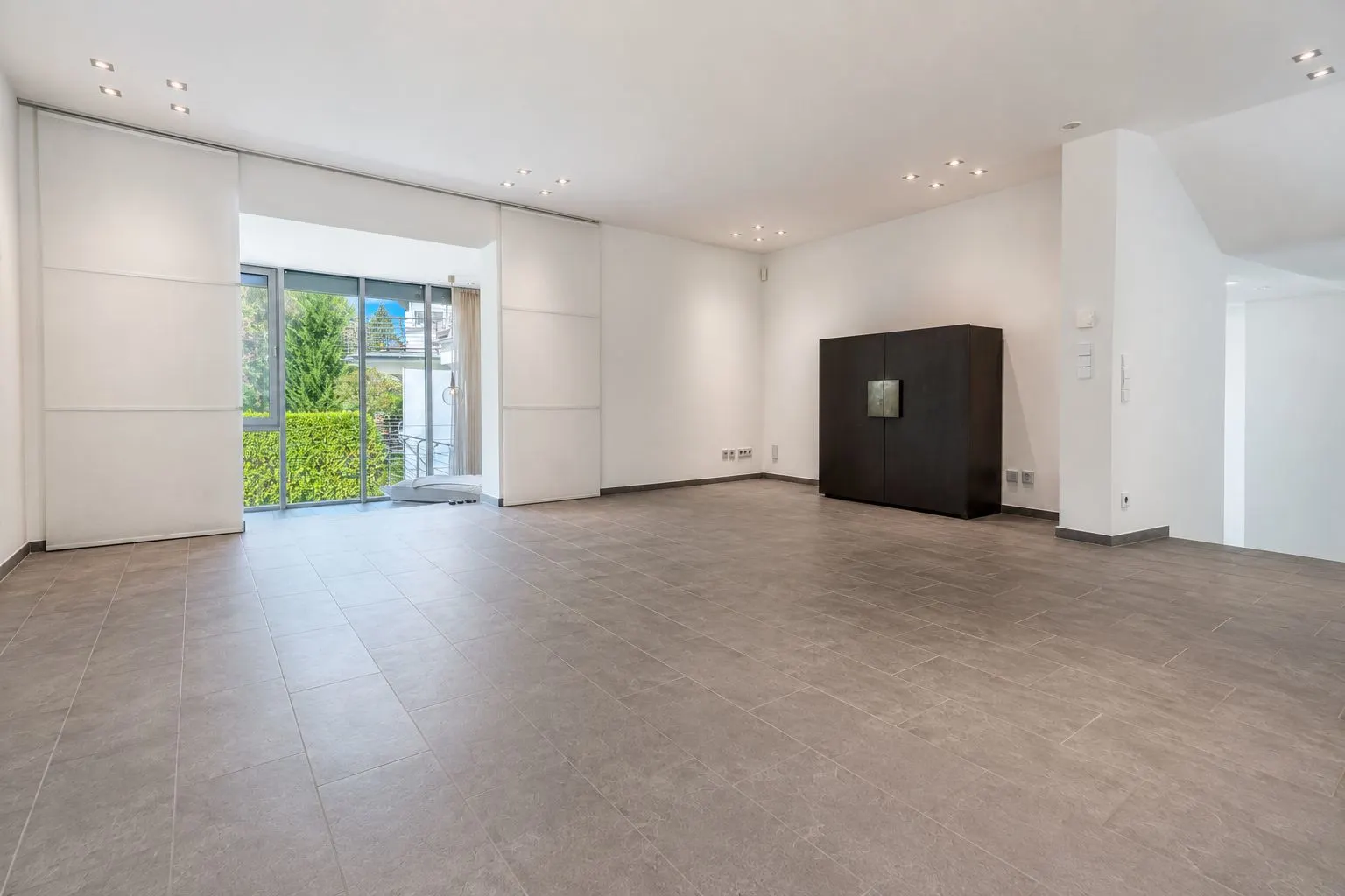 Großes, leeres Wohnzimmer mit Fliesenboden, weißen Wänden und großem Fenster zur Terrasse.