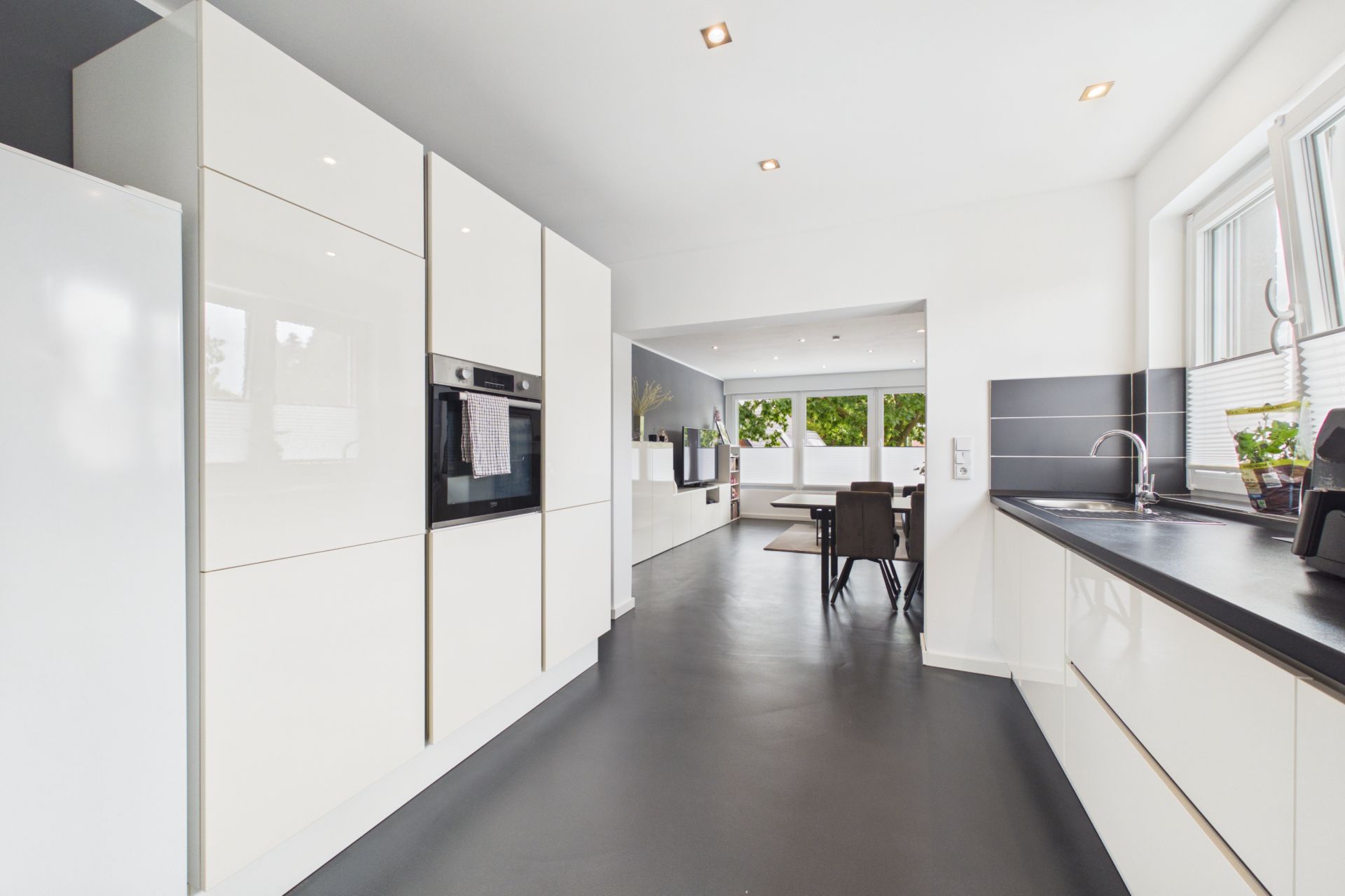 Moderne weiße Küche mit Einbauschränken, Ofen, Spüle und Blick auf Essbereich mit großen Fenstern.