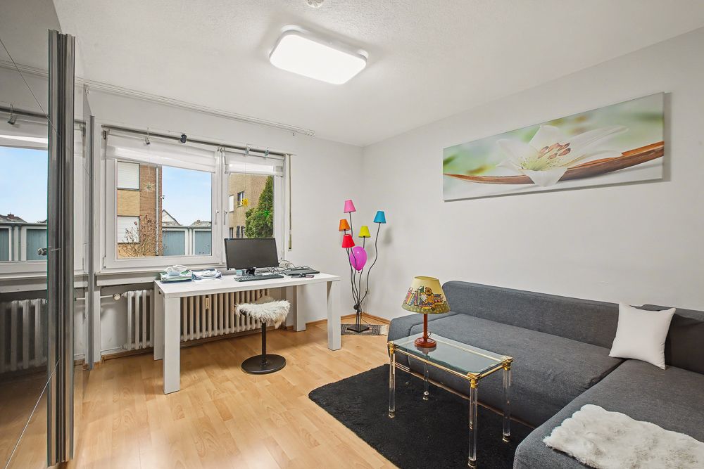 Helles Wohn- und Arbeitszimmer mit Schreibtisch, Computer, Sofa, Glastisch und bunter Stehlampe.