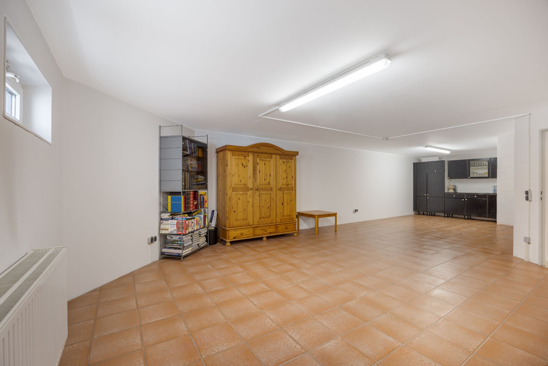 Großer Raum mit Fliesenboden, Holzschrank, Regal mit Büchern und Schränken in der Ecke.