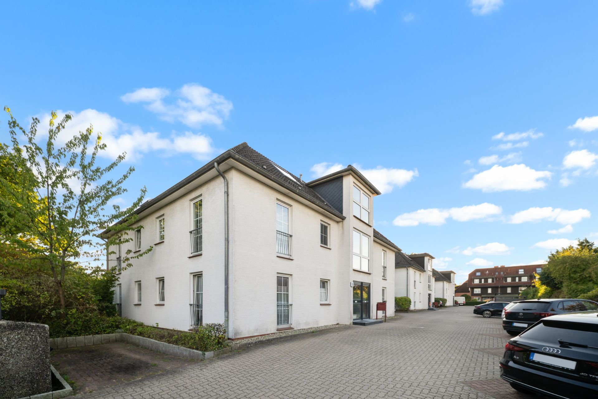 Weiße Wohngebäude mit mehreren Fenstern, davor gepflasterter Parkplatz mit geparkten Autos.