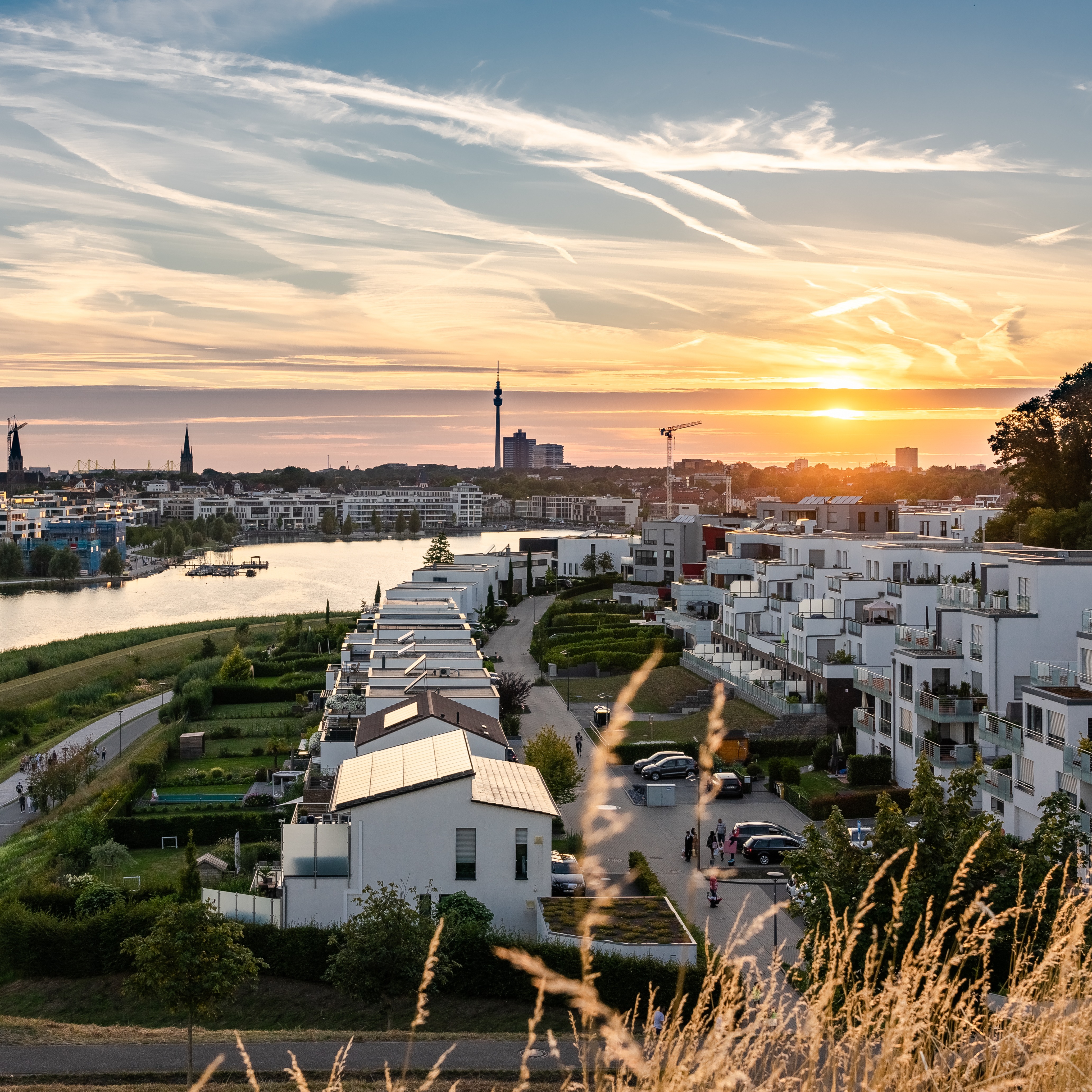 Moderne Wohngebäude am Flussufer bei Sonnenuntergang, mit Blick auf die Stadt im Hintergrund.