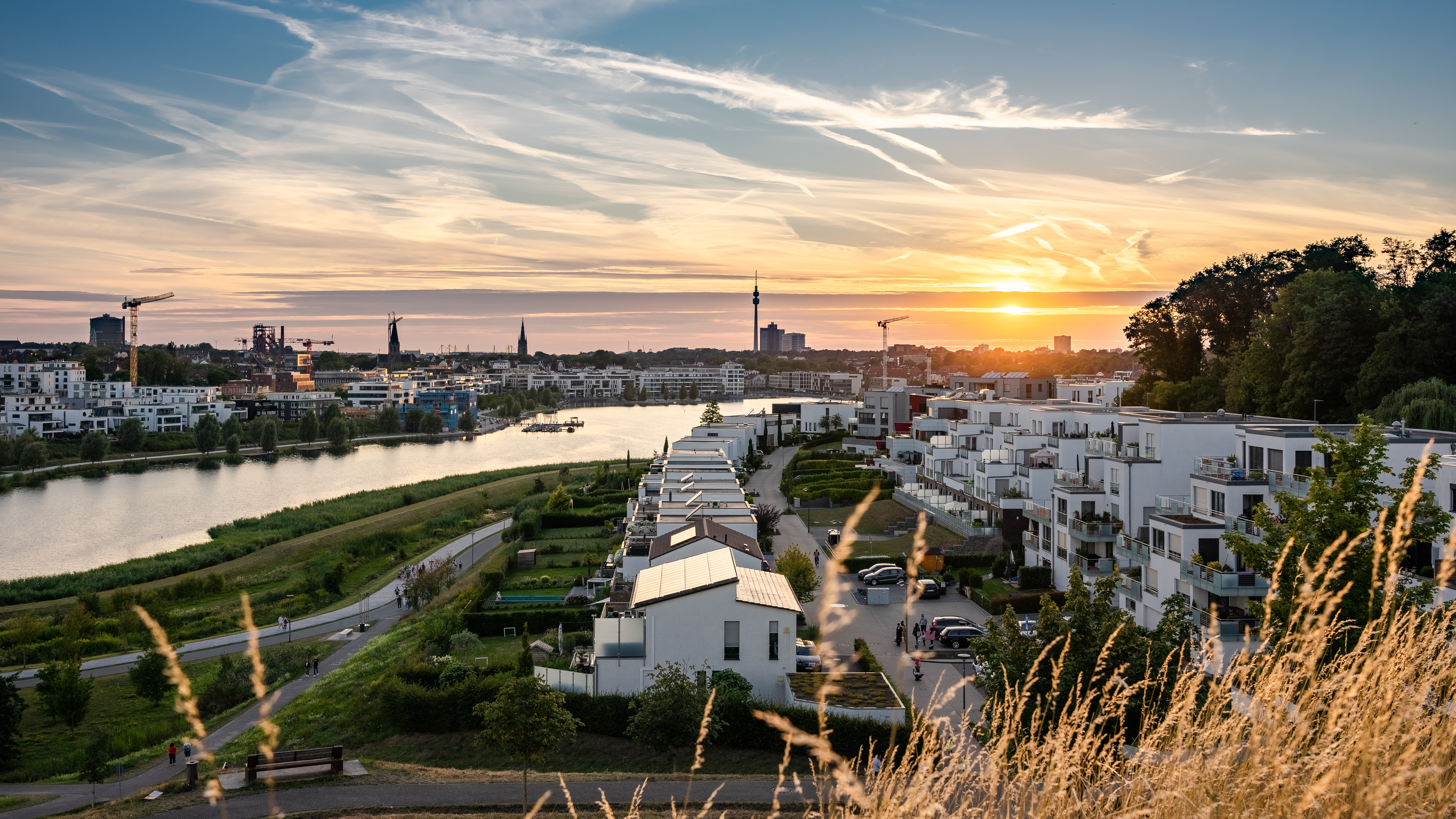 Moderne Wohngebäude am Flussufer bei Sonnenuntergang, mit Blick auf die Stadt im Hintergrund.