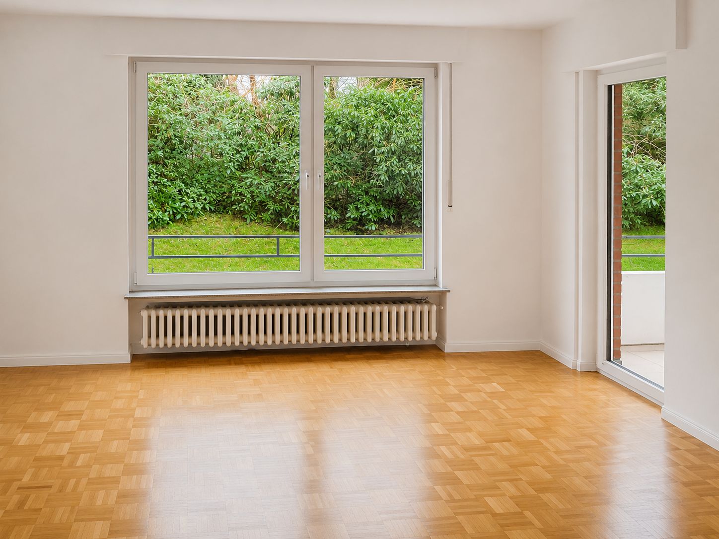 Leeres Zimmer mit Parkettboden, großem Fenster, Heizung und Zugang zur Terrasse, Blick auf grünen Garten.