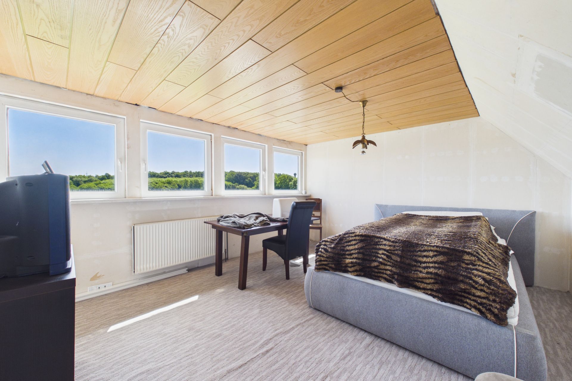 Helles Schlafzimmer mit großem Bett, Schreibtisch, Stuhl und mehreren Fenstern mit Blick ins Grüne.