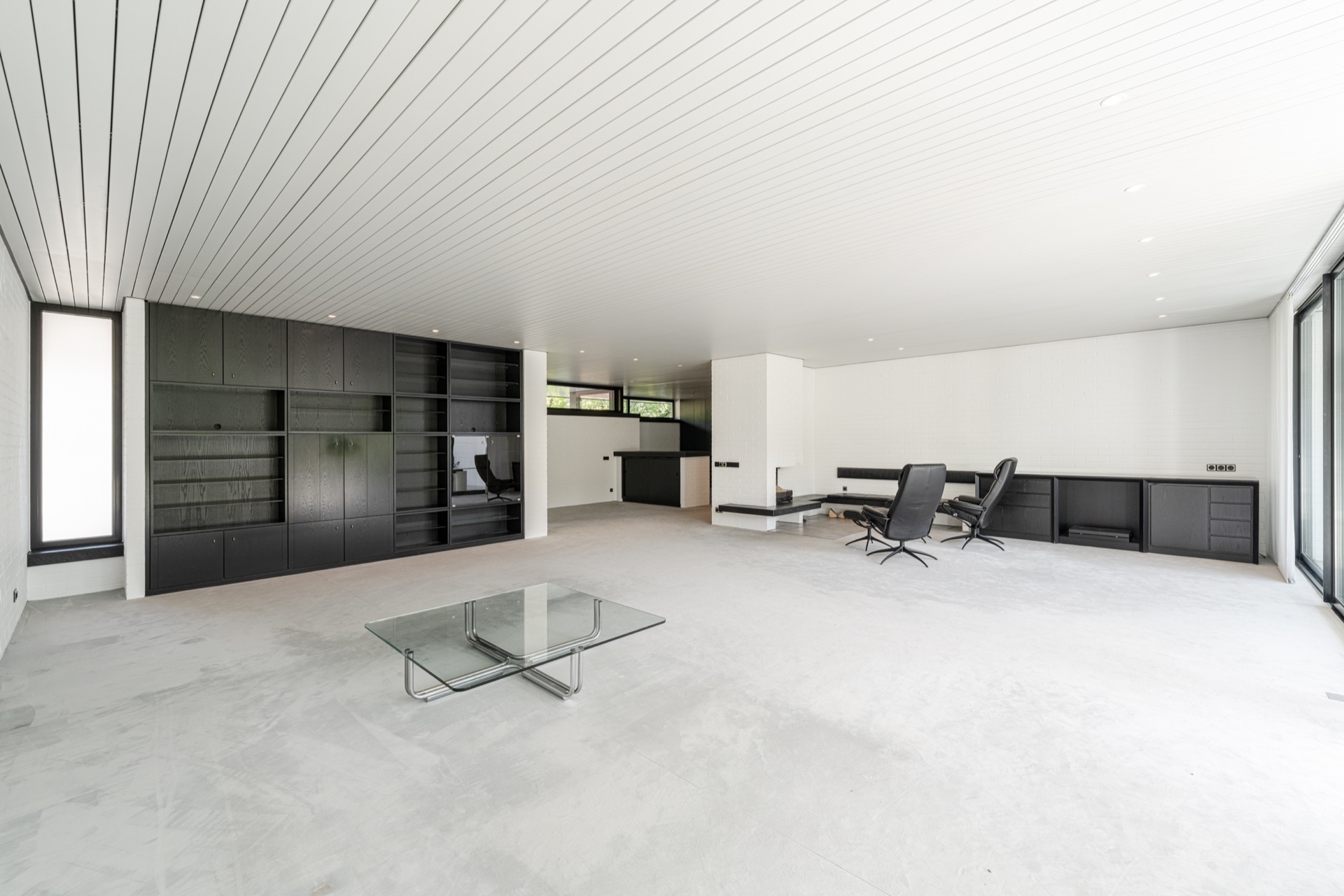 Heller, moderner Wohnraum mit Glastisch, schwarzen Regalen und Schreibtischen an der Wand.