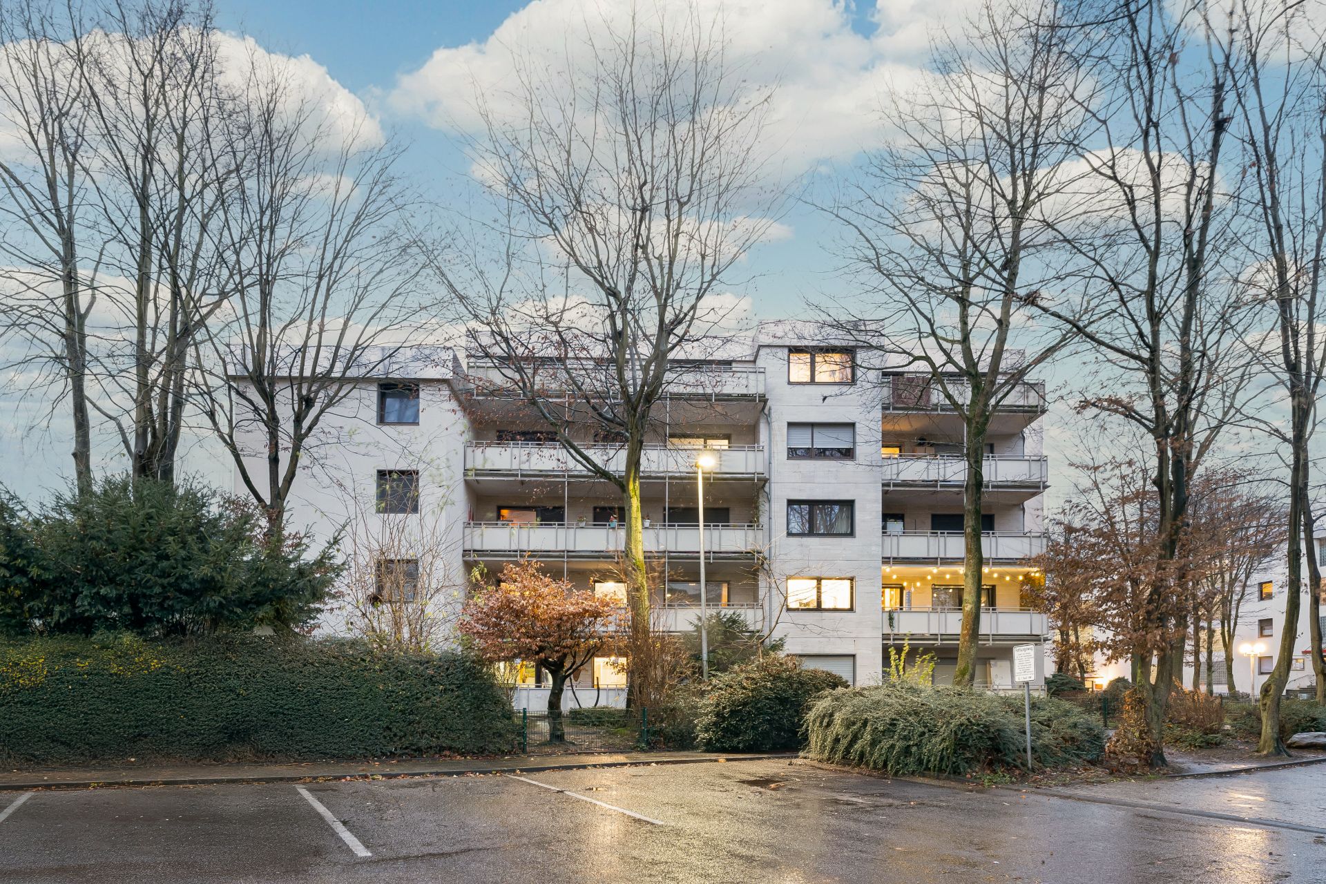 Wohngebäude mit mehreren Balkonen, Bäumen im Vordergrund und leerem Parkplatz.
