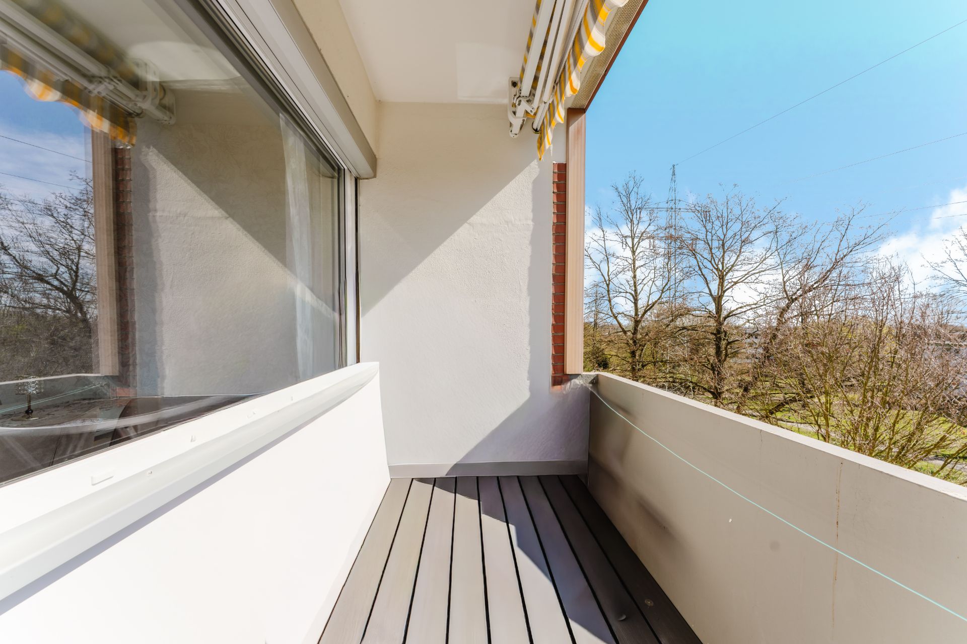 Heller Balkon mit Markise, Holzboden und Blick auf Bäume.