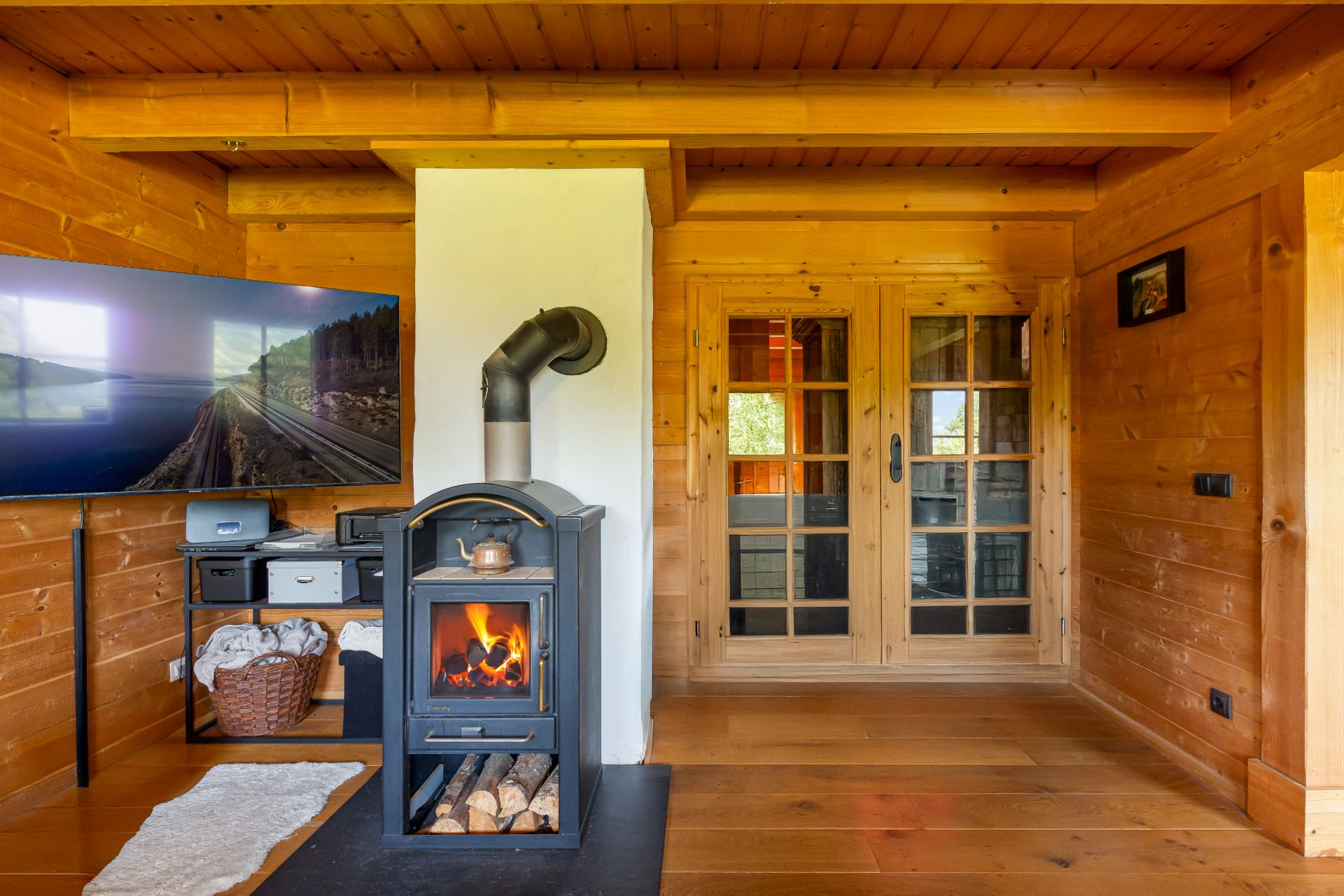 Wohnzimmer mit Holzofen, Fernseher, Holzwänden und Doppeltür aus Glas.