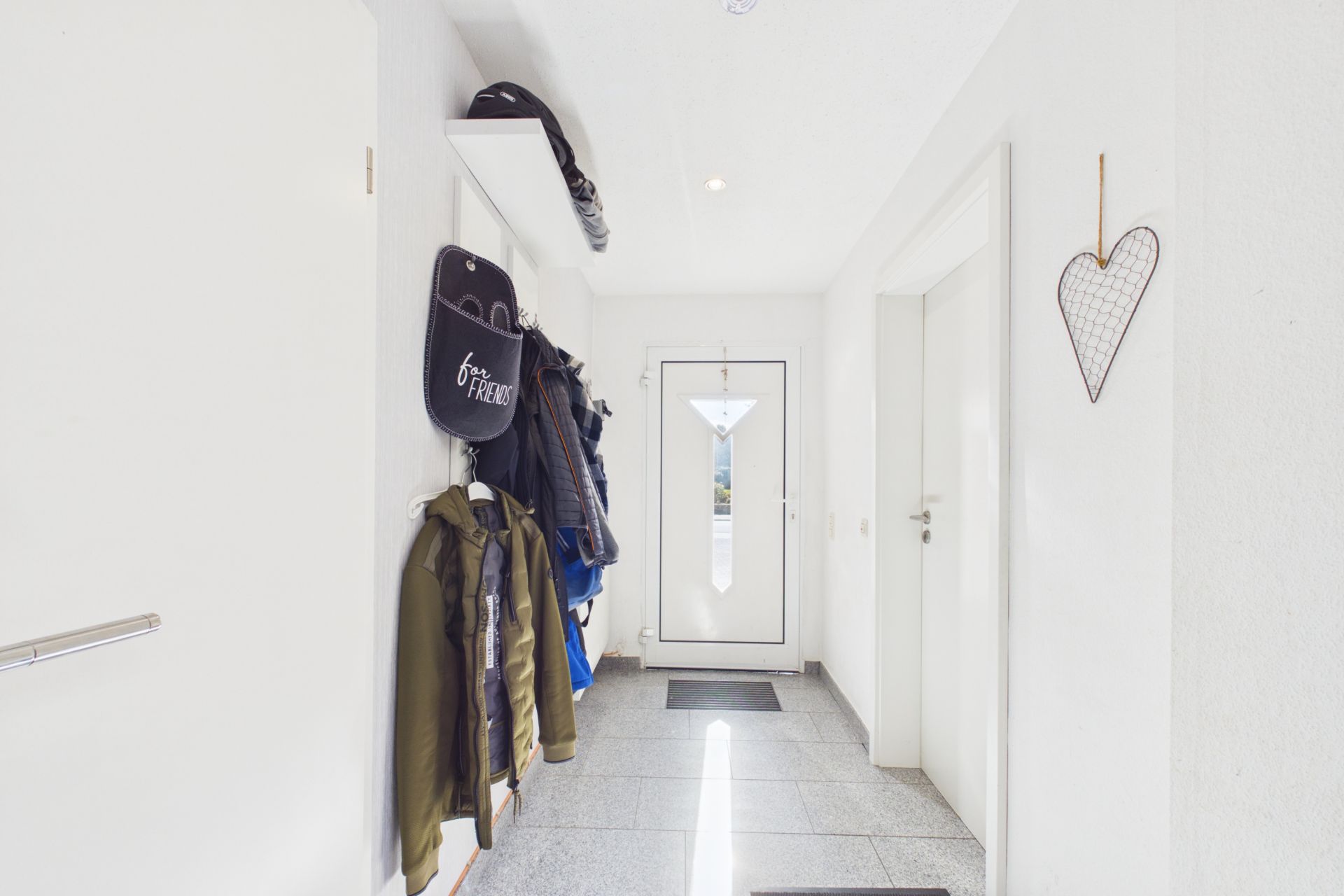 Heller Flur mit Garderobe, Jacken, Haken und einer weißen Haustür mit Glaseinsatz.