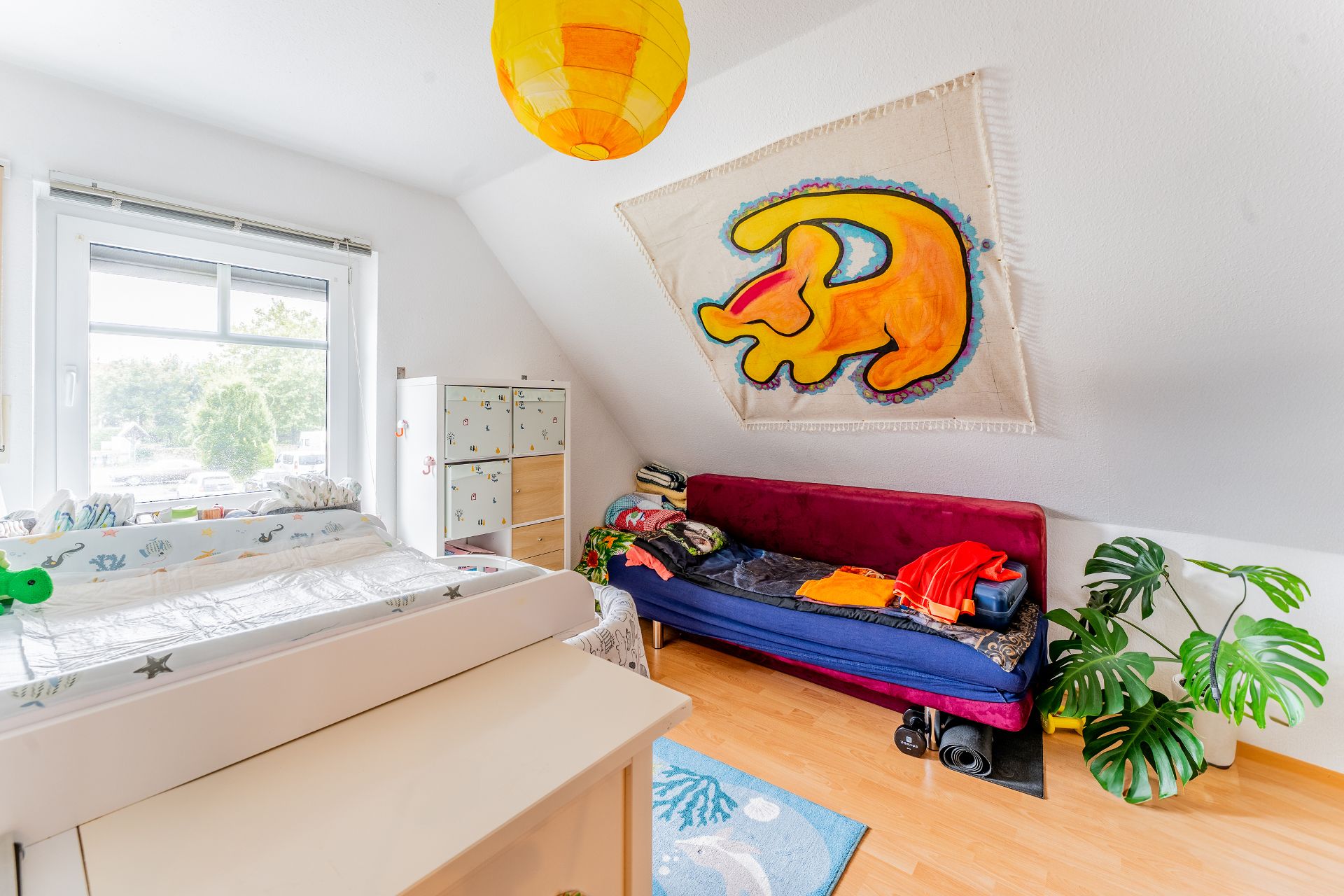 Helles Kinderzimmer mit Wickeltisch, Sofa, Pflanze und buntem Wandteppich.
