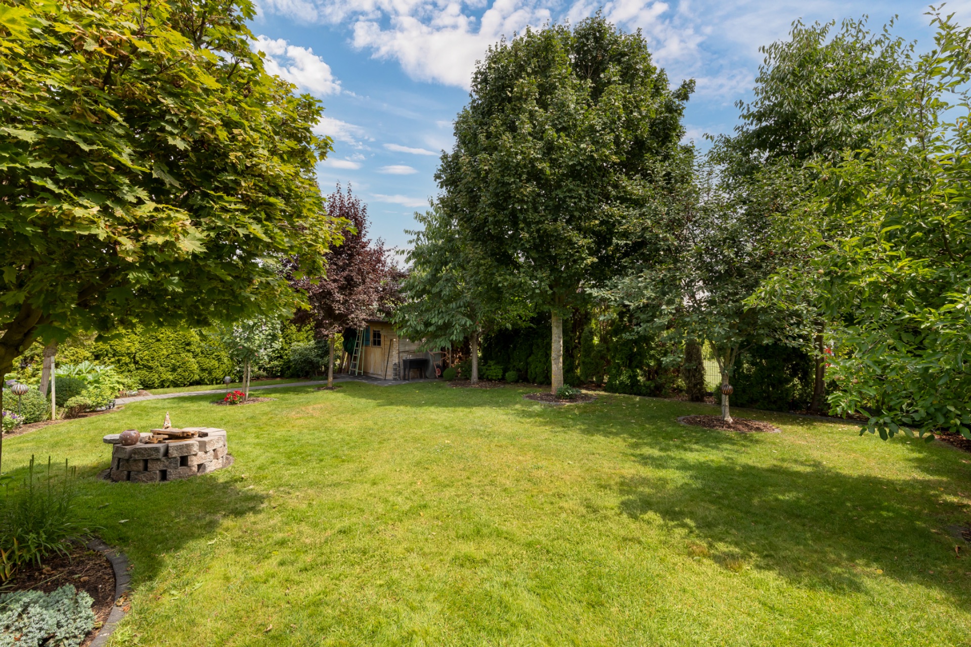 Großer Garten mit grünem Rasen, mehreren Bäumen und einer Feuerstelle aus Steinen.