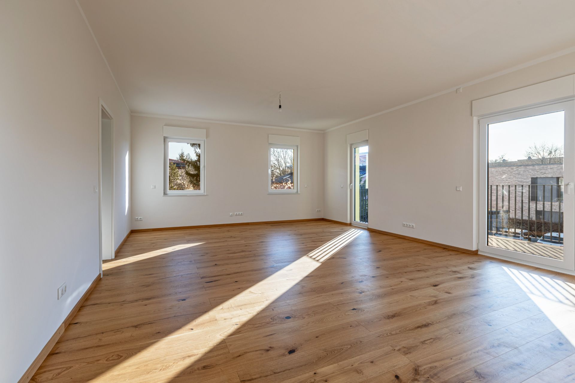Helles, leeres Zimmer mit Holzfußboden, zwei Fenstern und zwei Glastüren zur Terrasse.