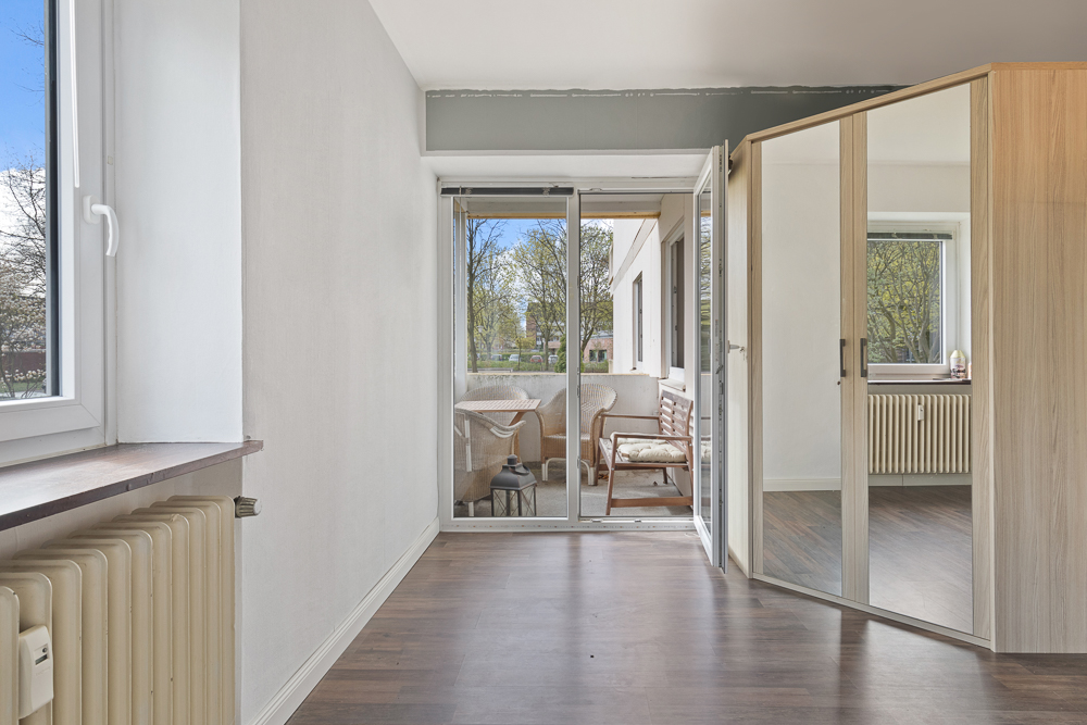 Helles Zimmer mit großem Spiegel, Holzfußboden und Zugang zu einem verglasten Balkon mit Sitzmöbeln.