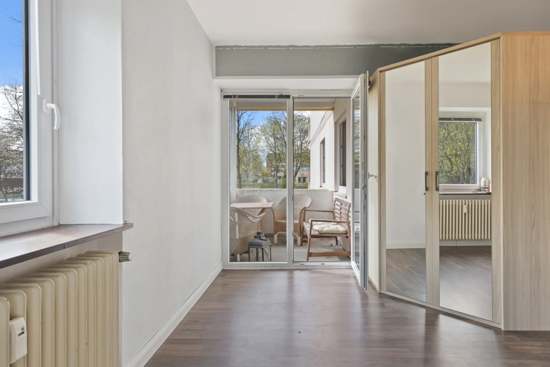 Helles Zimmer mit großem Spiegel, Holzfußboden und Zugang zu einem verglasten Balkon mit Sitzmöbeln.