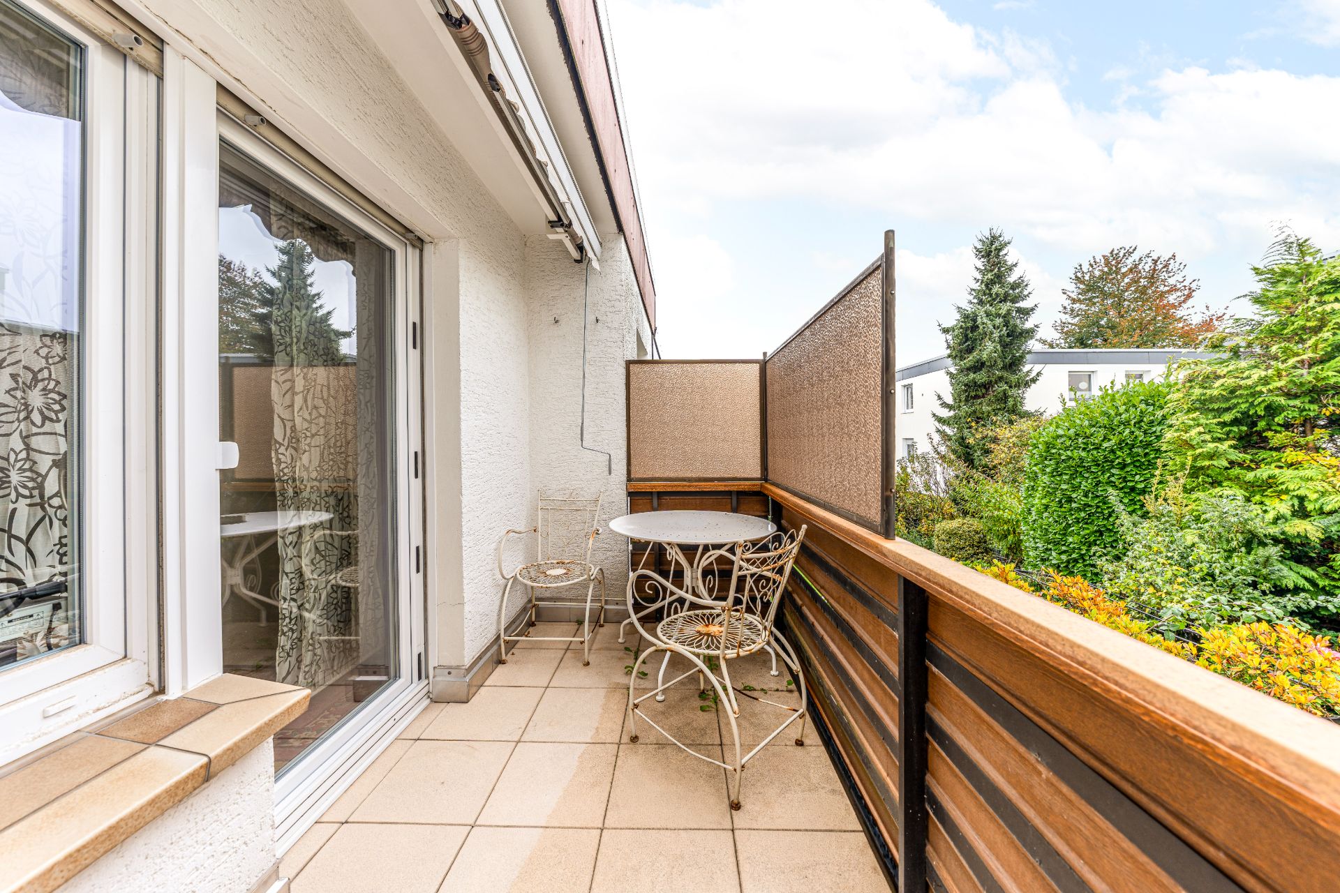 Balkon mit zwei Stühlen und einem runden Tisch, daneben Fenster und Blick auf Garten.