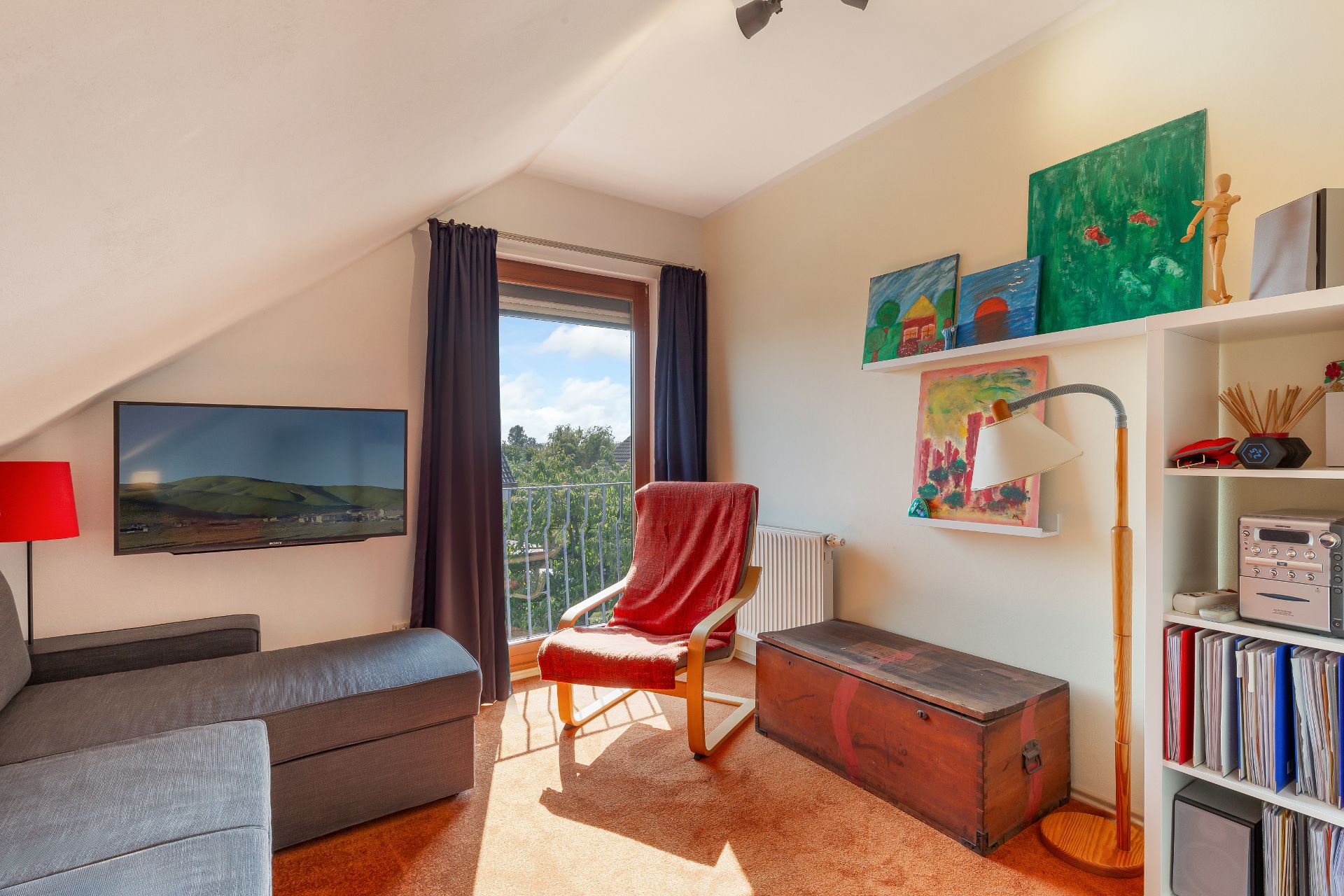 Helles Wohnzimmer mit grauem Sofa, rotem Sessel, Wandregal, Bildern und Blick nach draußen.