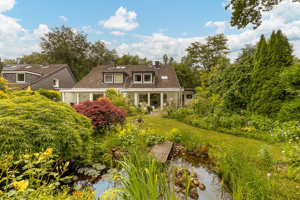 Haus mit großem Garten, Teich und vielen Pflanzen im Vordergrund, bei Tageslicht aufgenommen.